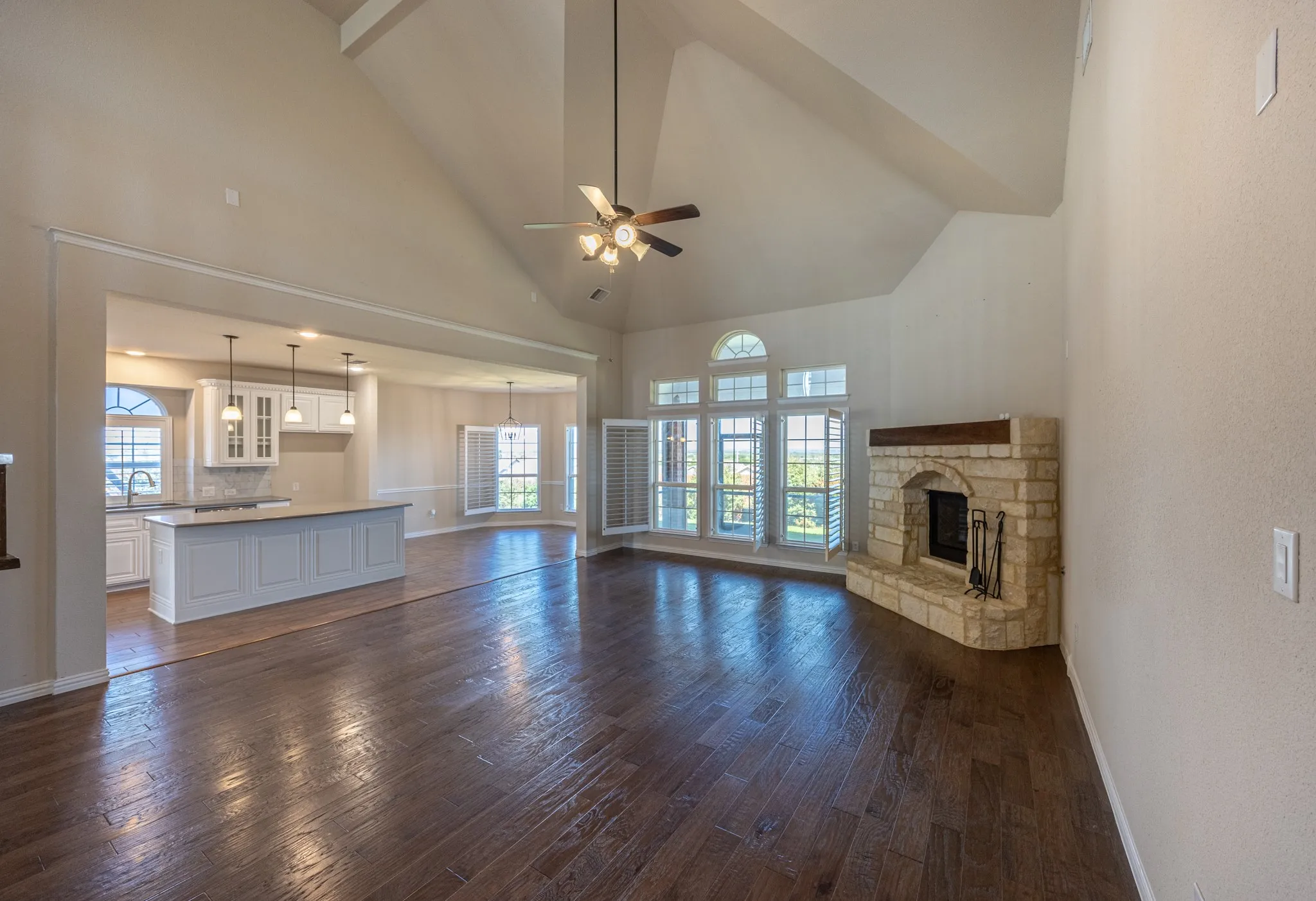 Vaulted ceilings accentuate this expansive living area featuring wood-finish flooring, a stone fireplace with a wood mantel, and multiple windows with plantation shutters