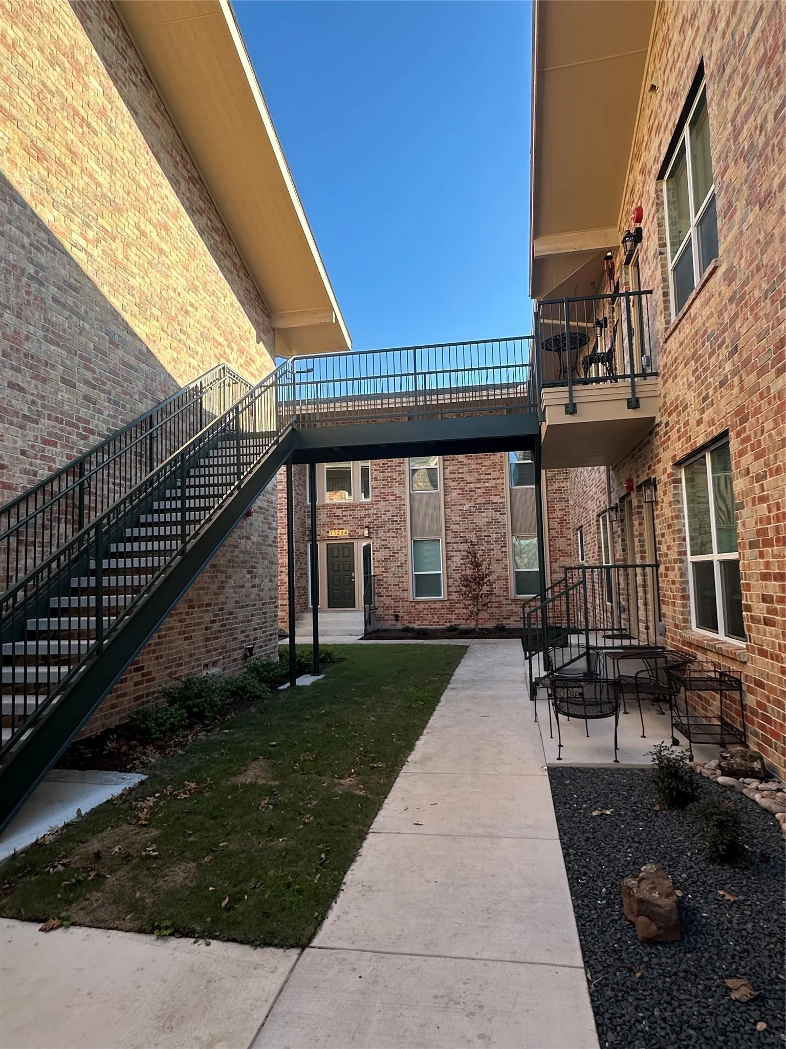 Brick exterior architecture featuring a second-story walkway with metal railings, a ground-level patio, and concrete pathways