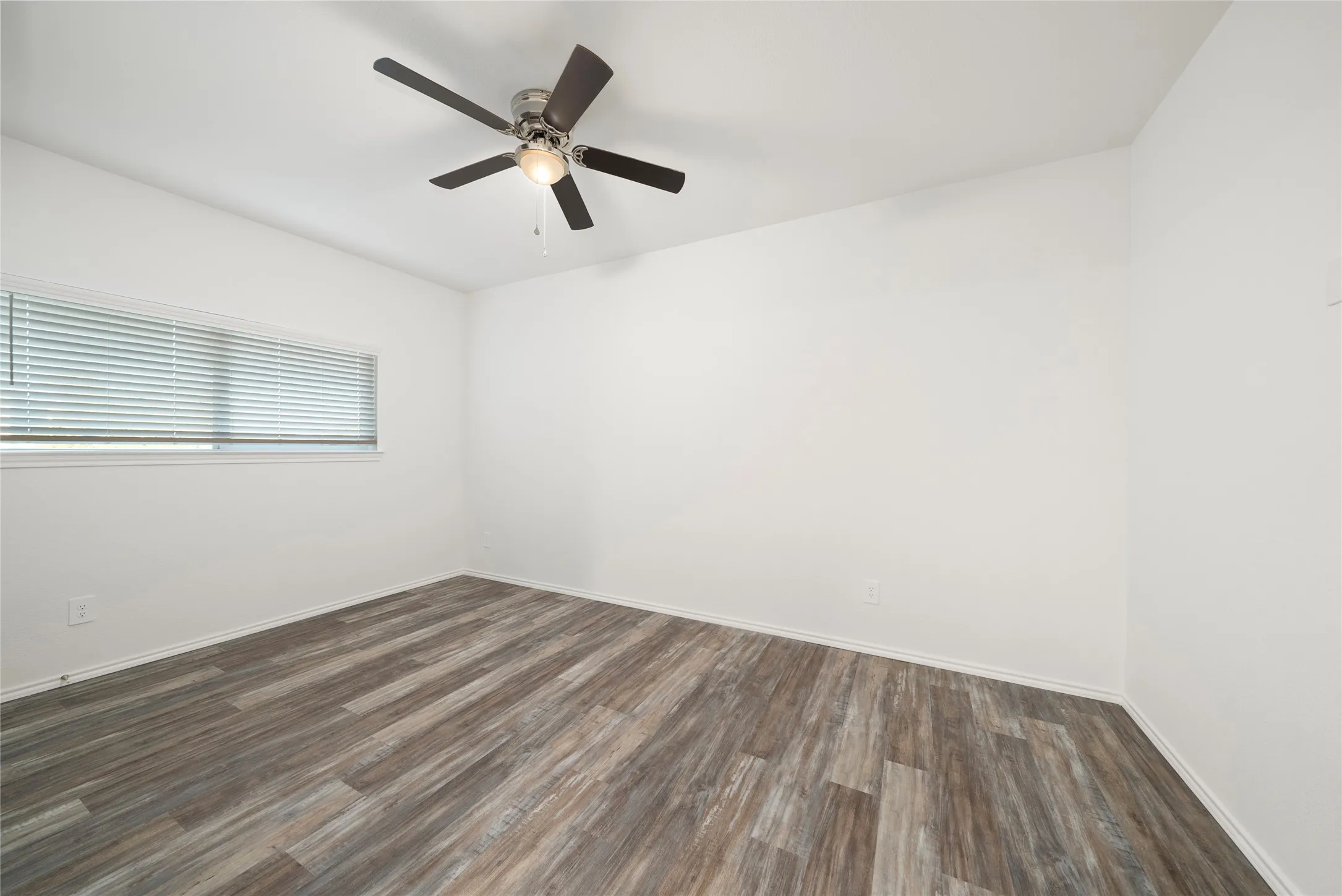 Spacious room featuring wood-finish flooring, a ceiling fan with integrated lighting, a window with horizontal blinds, and clean white walls