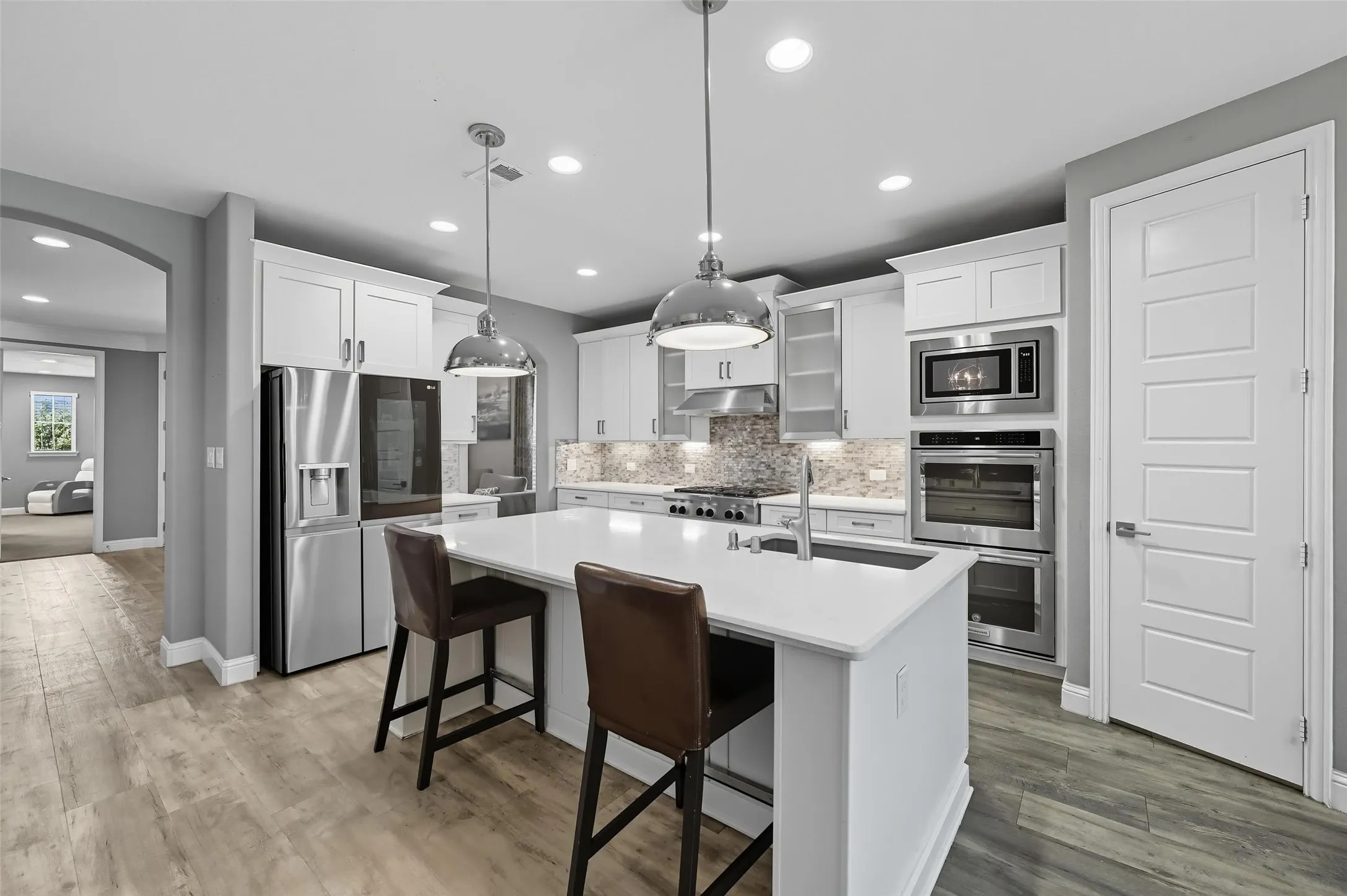 Modern kitchen featuring a large central island with seating, stainless steel appliances, light wood-finish flooring, white cabinetry, and a mosaic tile​​‌​​​​‌​​‌‌​‌‌​​​‌‌​‌​‌​‌​​​‌​​ backsplash