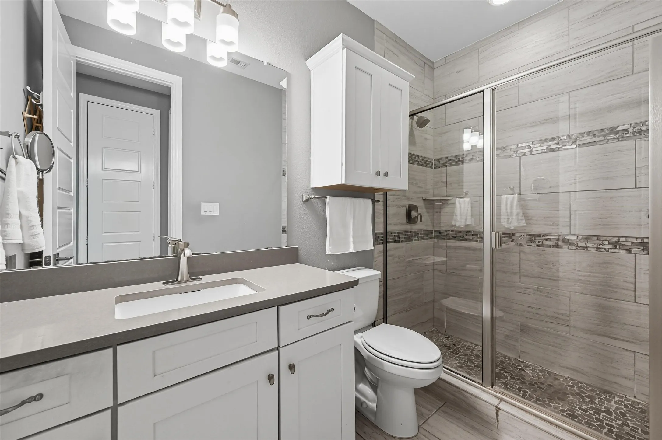 Contemporary bathroom featuring a spacious walk-in shower with tile surround and pebble flooring, a dual-sink vanity with light-toned countertops, and white cabinetry