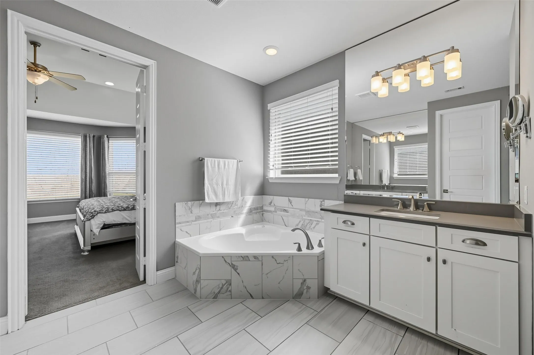 Bathroom featuring a corner soaking tub with marble-look tile surround, a single vanity with dark countertop and white cabinetry, and large format light-toned tile flooring