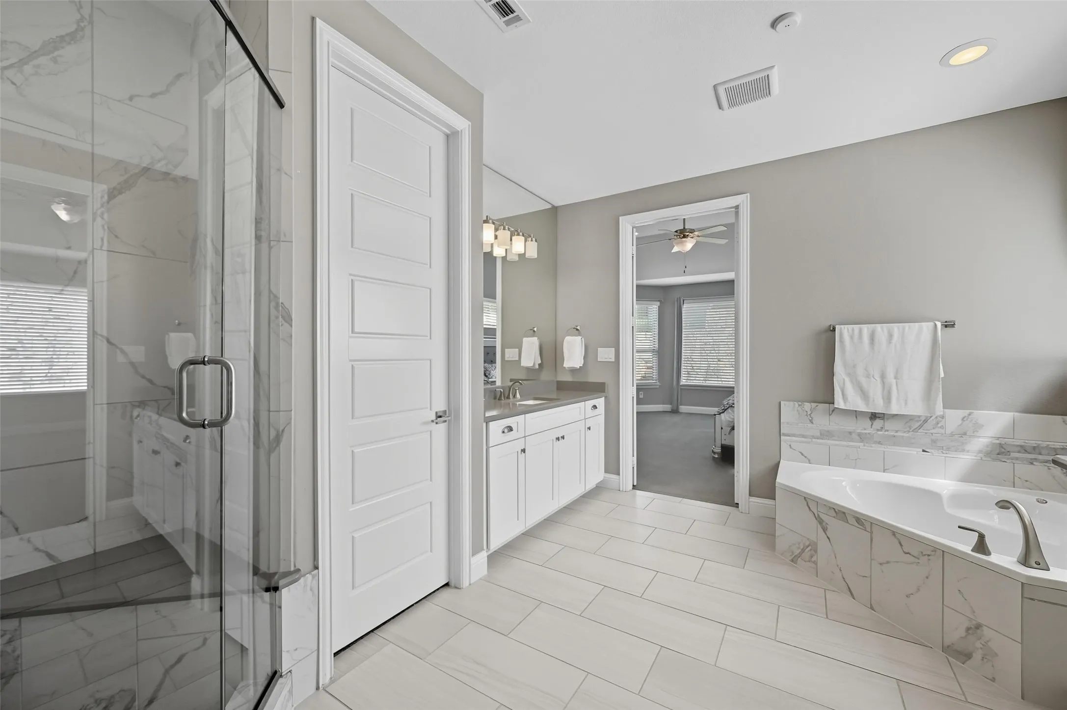 Spacious bathroom featuring a glass-enclosed shower with marble-look tile, a built-in jetted tub, and dual vanities with white cabinetry and stone countertops