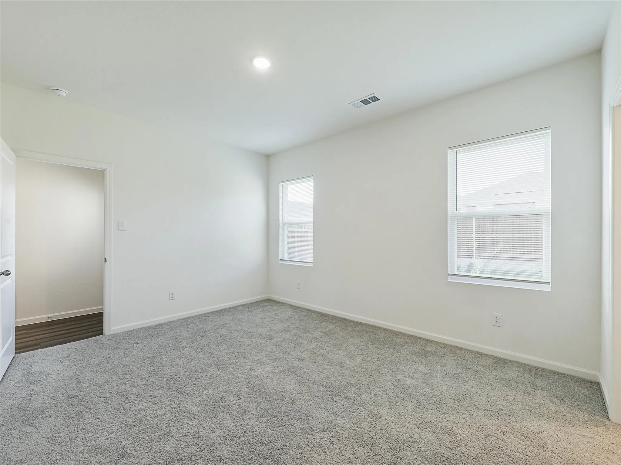 Spacious room featuring plush gray carpeting, white walls, two windows with blinds, a recessed ceiling light, and an open doorway revealing wood-finish flooring