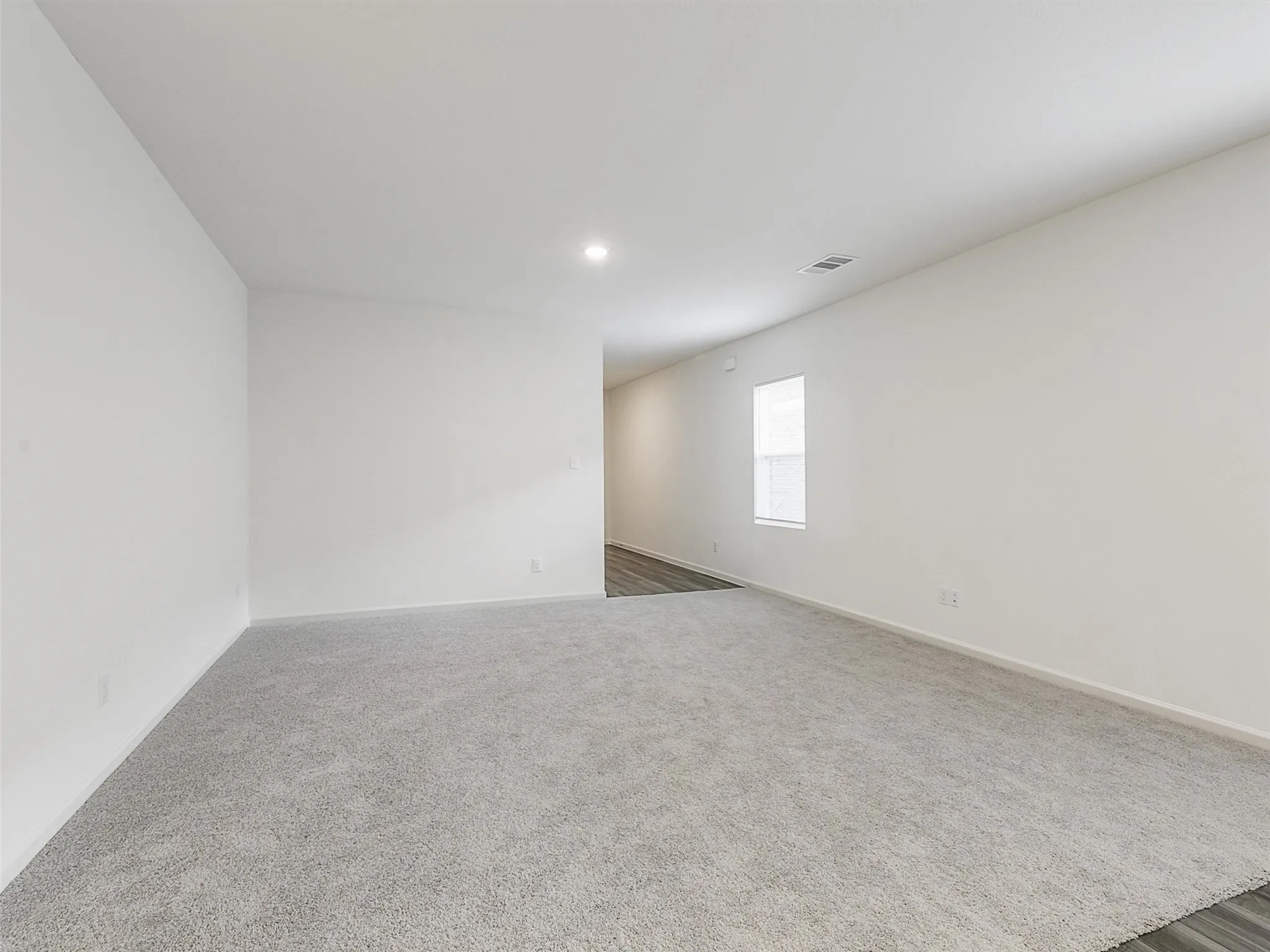 Spacious room featuring light gray carpeting, neutral wall finishes, a recessed ceiling light, and a single window allowing ambient illumination
