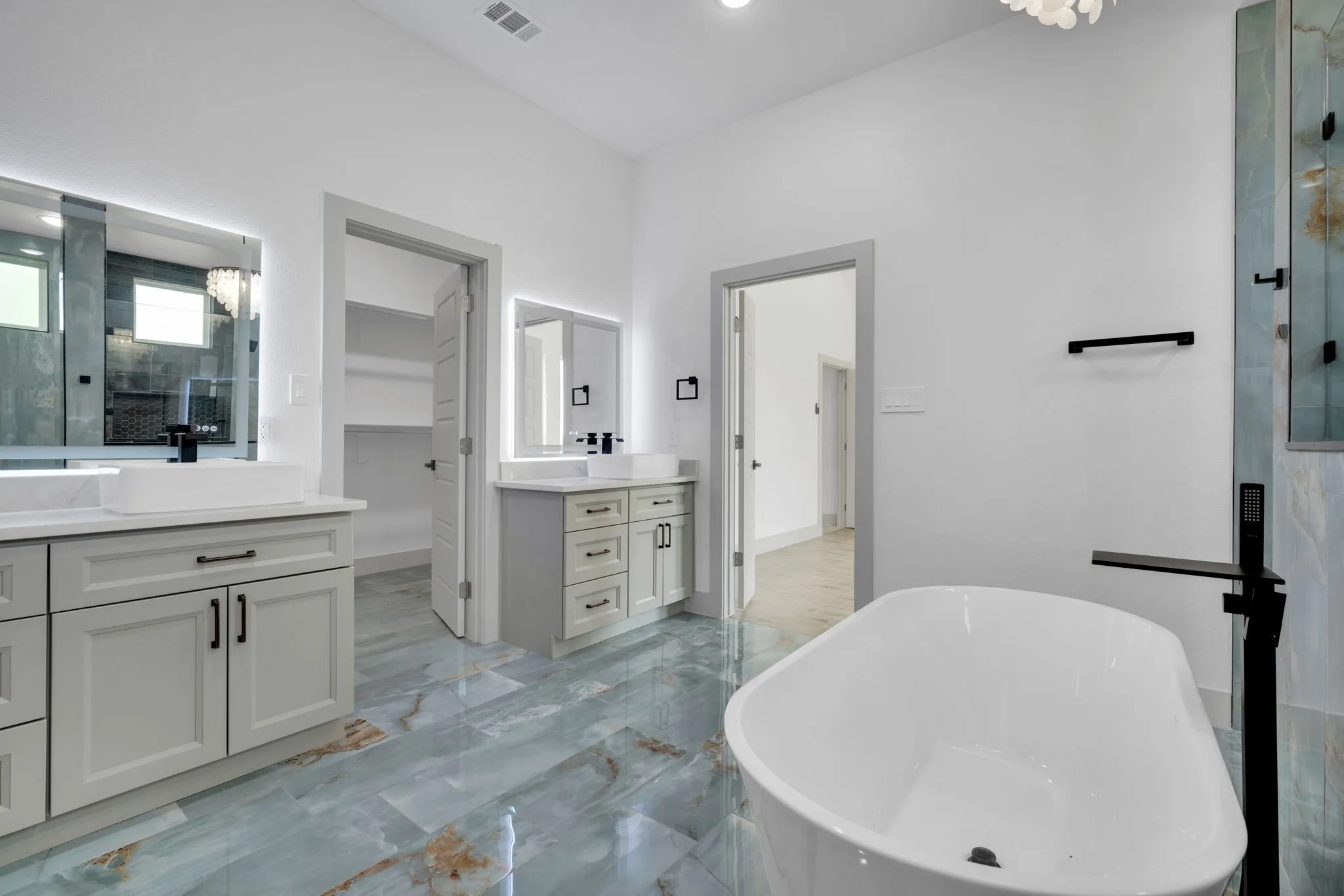 Luxurious primary bathroom featuring a freestanding soaking tub with matte black fixtures, dual vanities with soft-close cabinetry and integrated lighting, large-format tile flooring with natural stone veining, frameless glass shower enclosure, and recessed ceiling lighting throughout