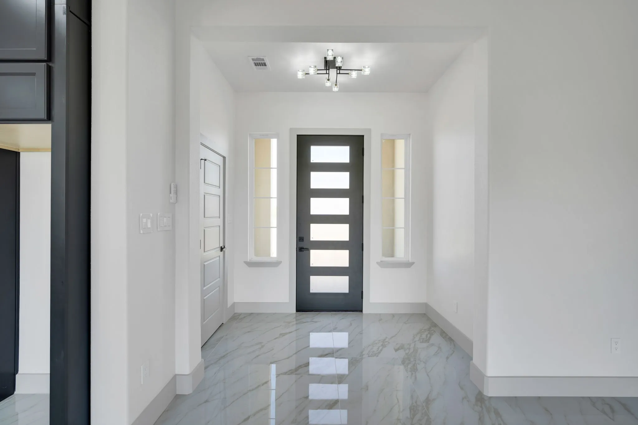 Bright entryway showcasing polished stone flooring, a modern black front door with horizontal glass panels and sidelights, recessed ceiling lighting, and clean white walls throughout