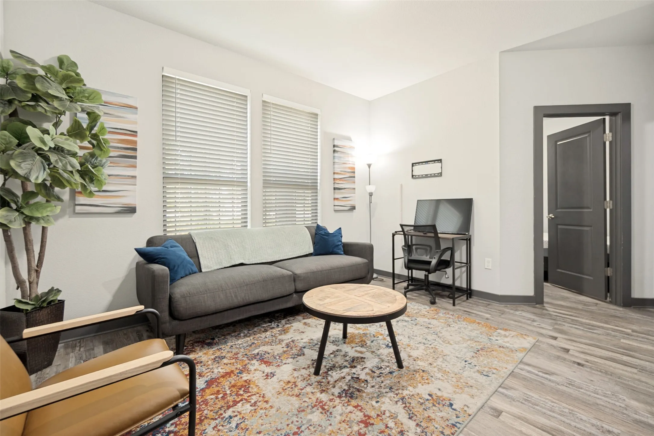 Bright, open living space showcasing wood-finish flooring, a charcoal upholstered sofa with blue accent pillows, round wood-top side table, and abundant natural light from large horizontal windows
