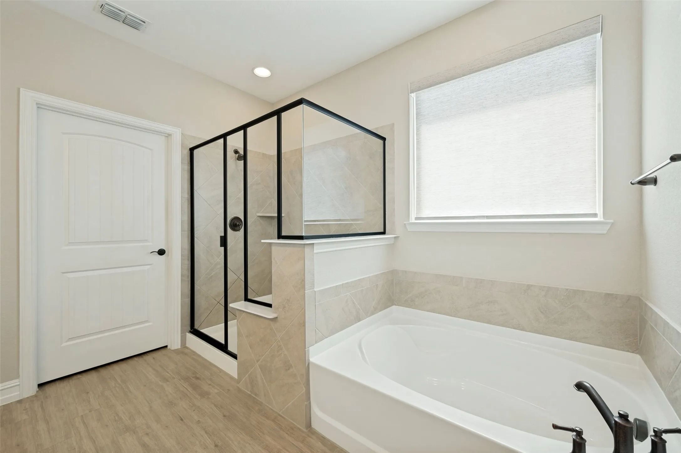 Spa-inspired primary bathroom featuring a freestanding soaking tub, walk-in shower enclosure with black metal framing and glass panels, neutral tile surround, wood-finish flooring, and abundant natural light from a large window