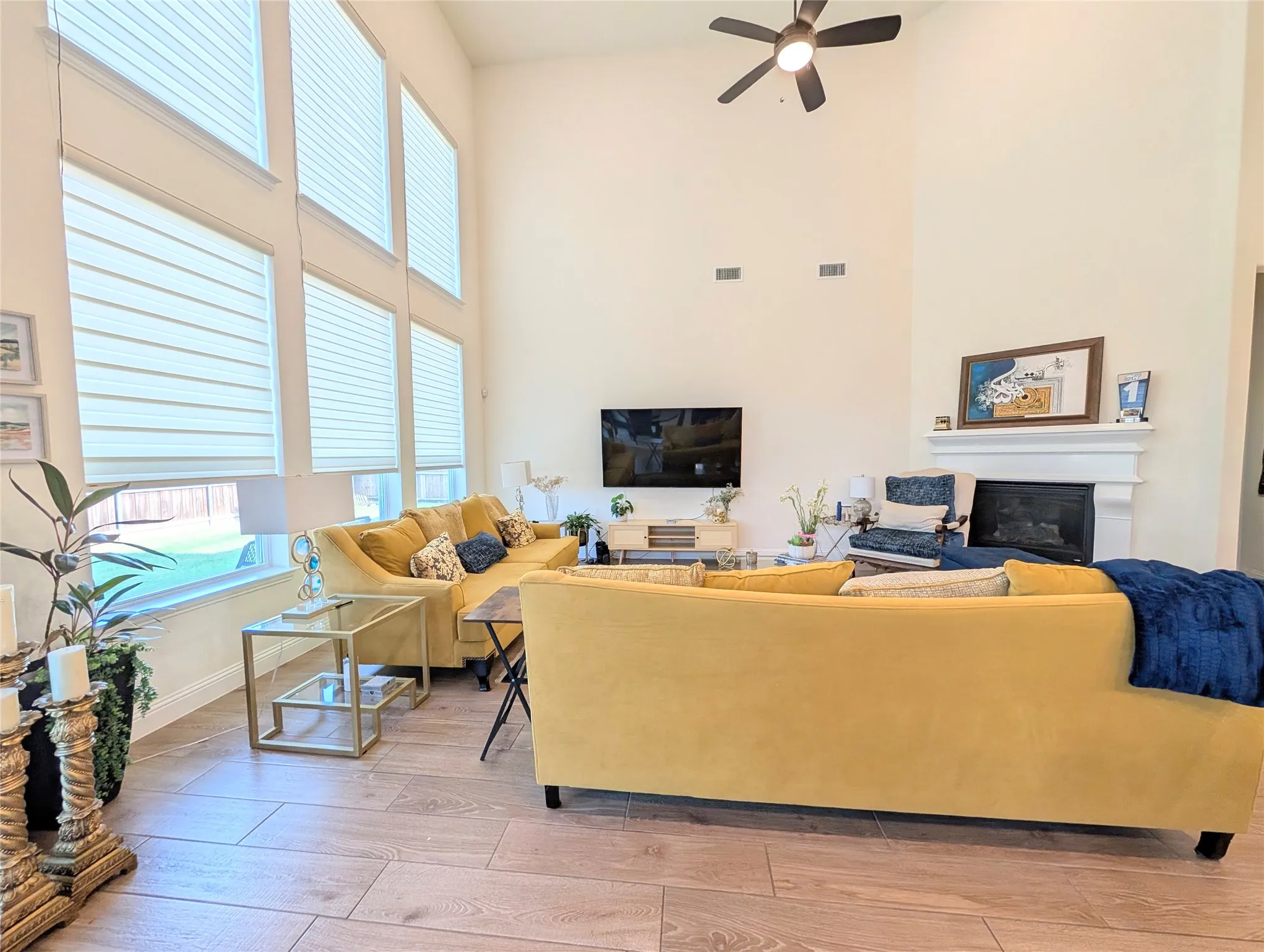 Spacious living area featuring high ceilings, tall windows with blinds, a ceiling fan, a fireplace with a mantel, and wood-finish flooring