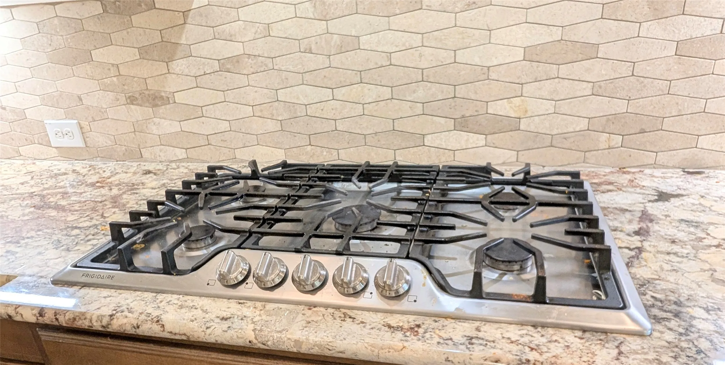 Stainless steel gas cooktop set into a variegated countertop, complemented by an elongated hexagonal tile backsplash