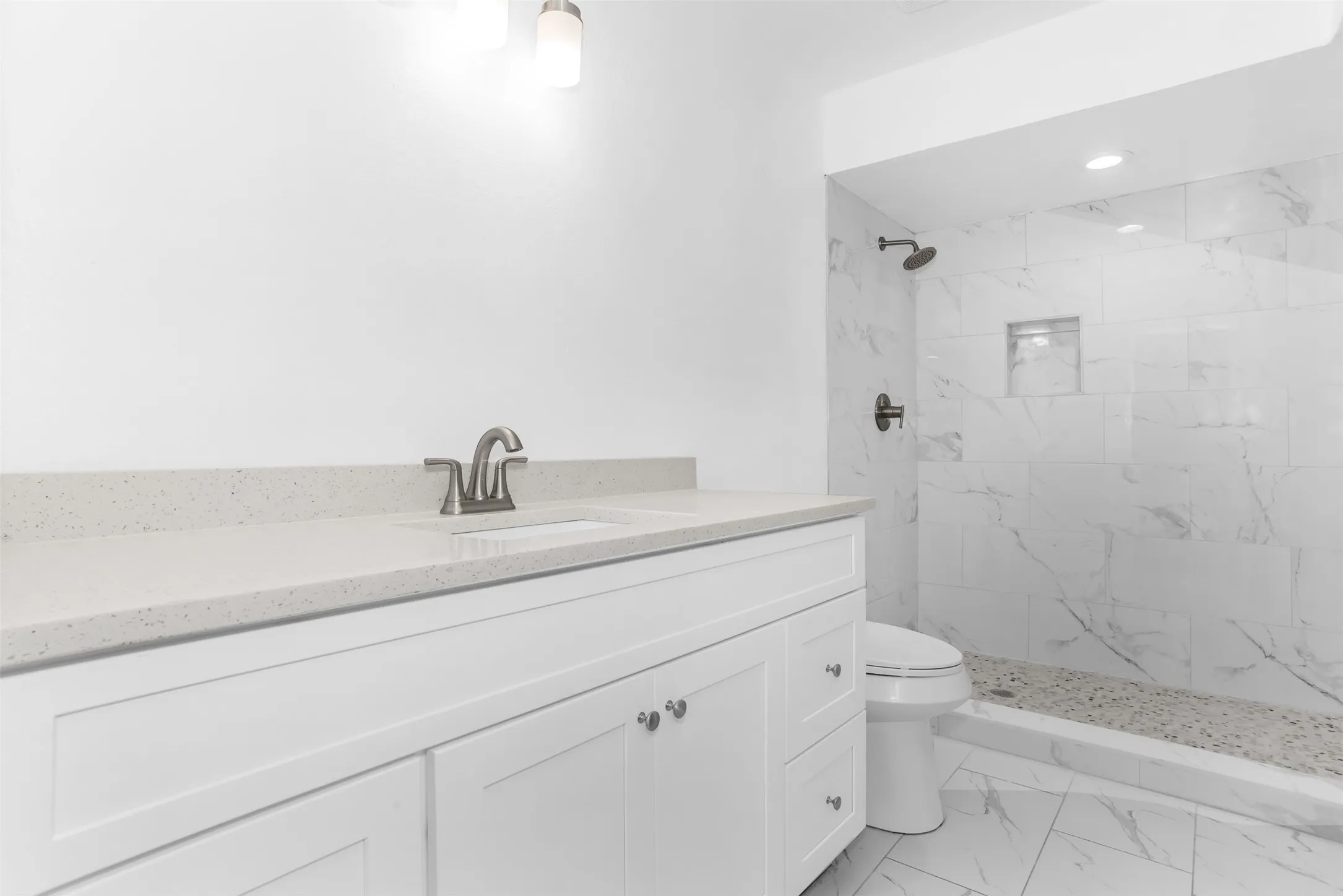 White shaker vanity with a light-colored countertop, brushed nickel faucet, and a tiled shower with a built-in niche