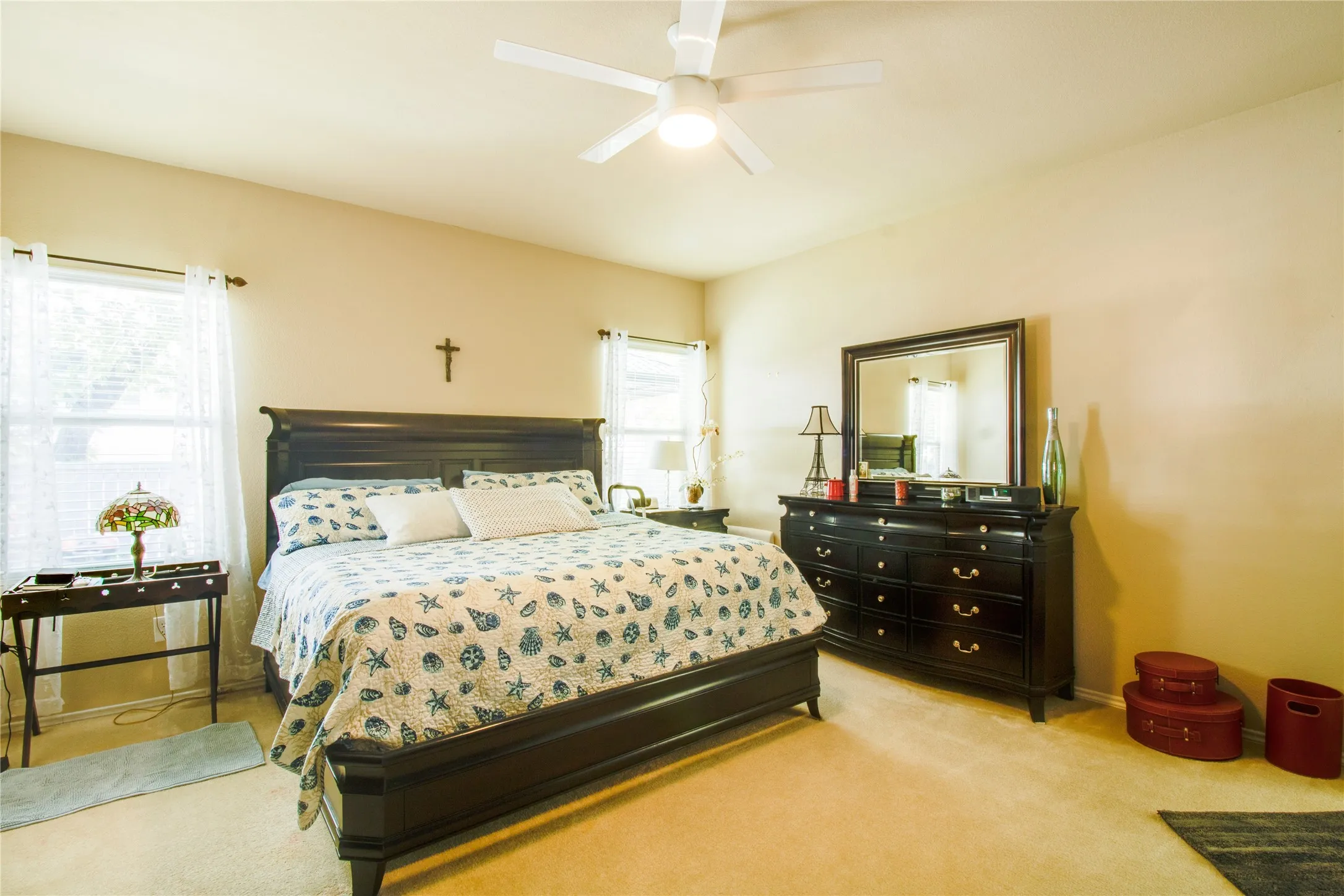 Carpeted bedroom featuring two windows with white sheer curtains, a white ceiling fan with integrated lighting, and neutral-toned walls