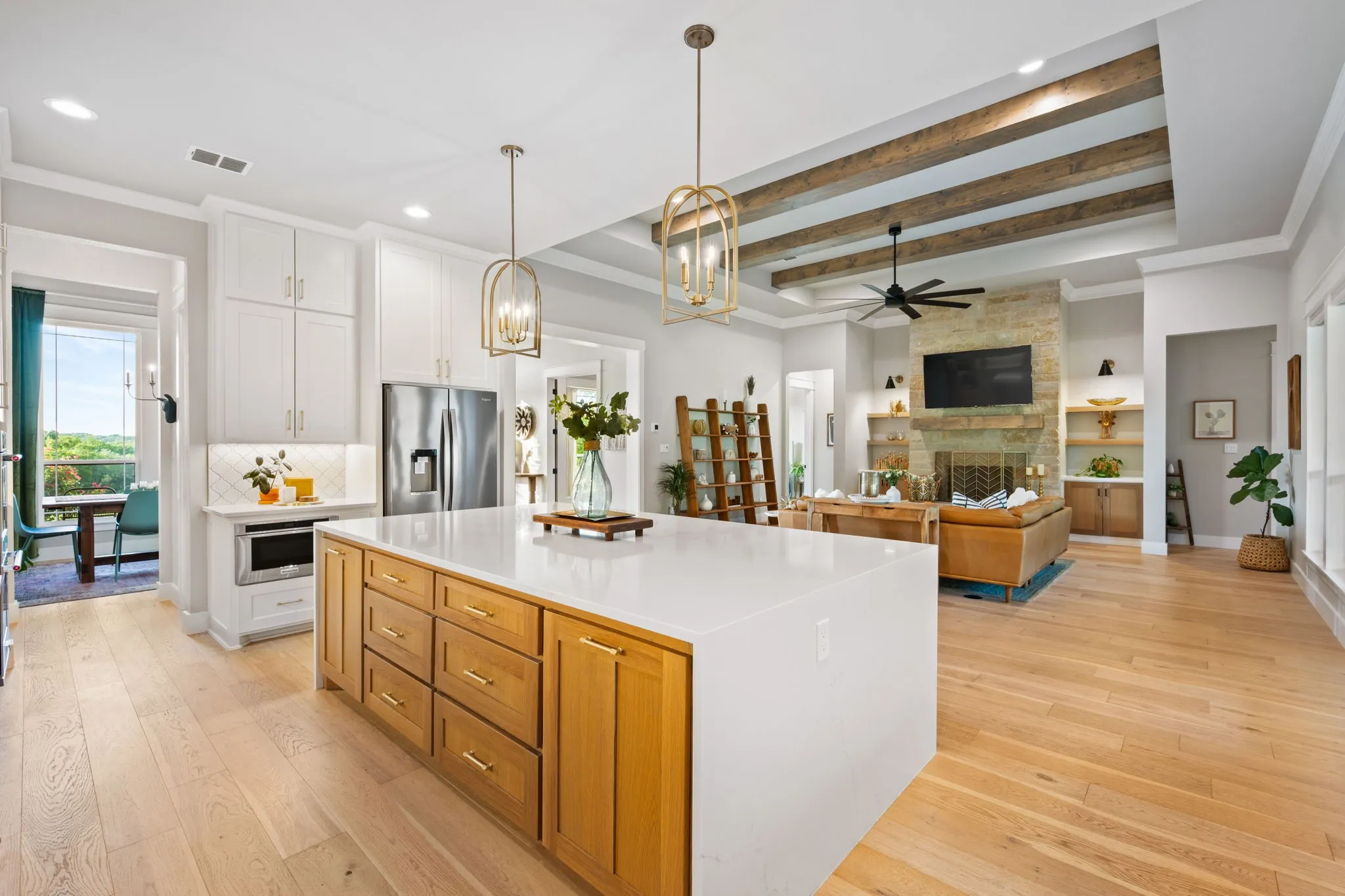 Open-concept kitchen and living area featuring a large white quartz island with wood-finish cabinetry base, stainless steel appliances, white shaker-style cabinetry, coffered ceiling with wood beam detailing, pendant lighting, ceiling fan, and a fireplace with built-in shelving visible in the adjacent living space