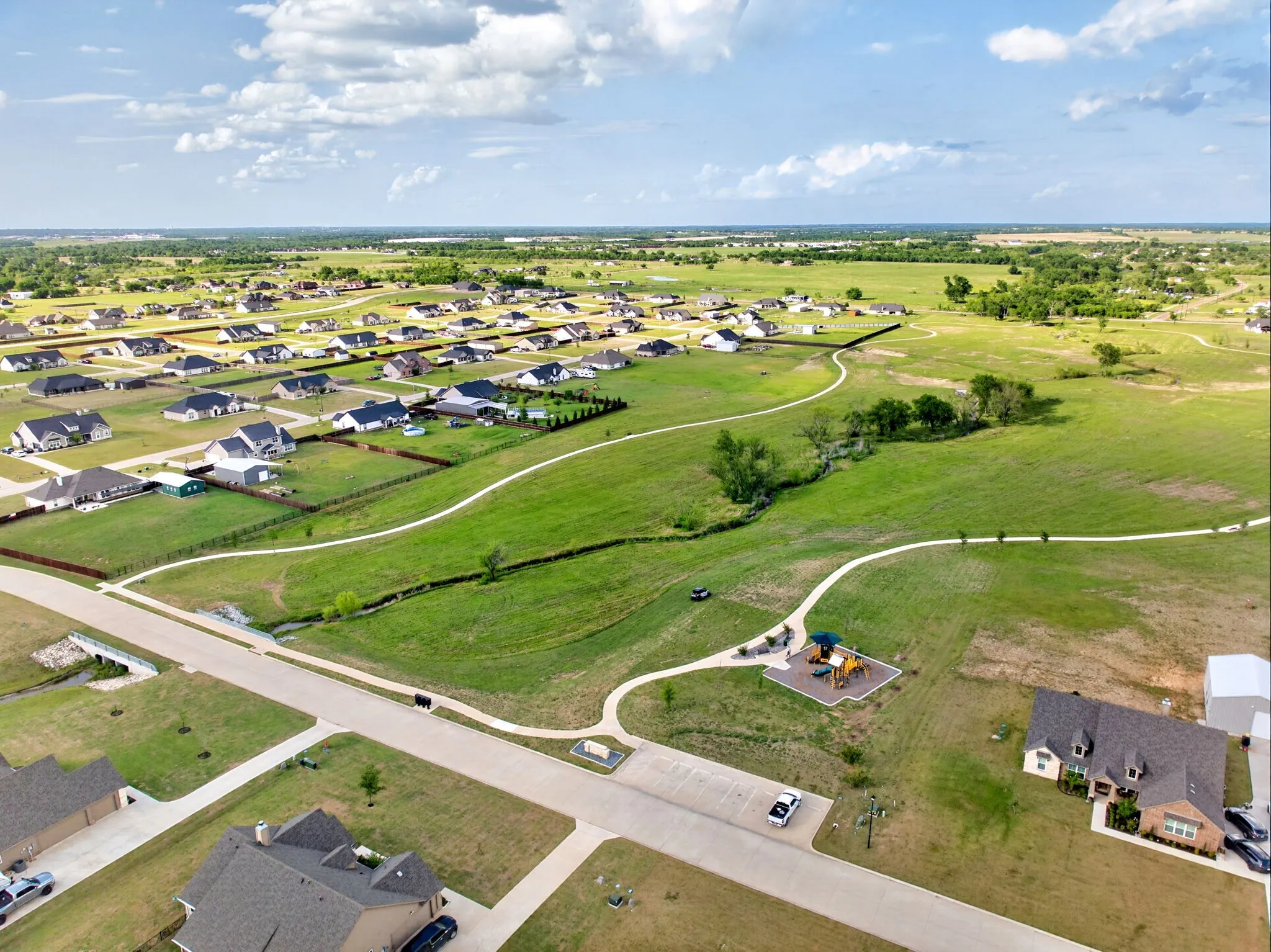 Expansive aerial capturing a developing neighborhood with residential properties, green spaces, walking paths, and a playground