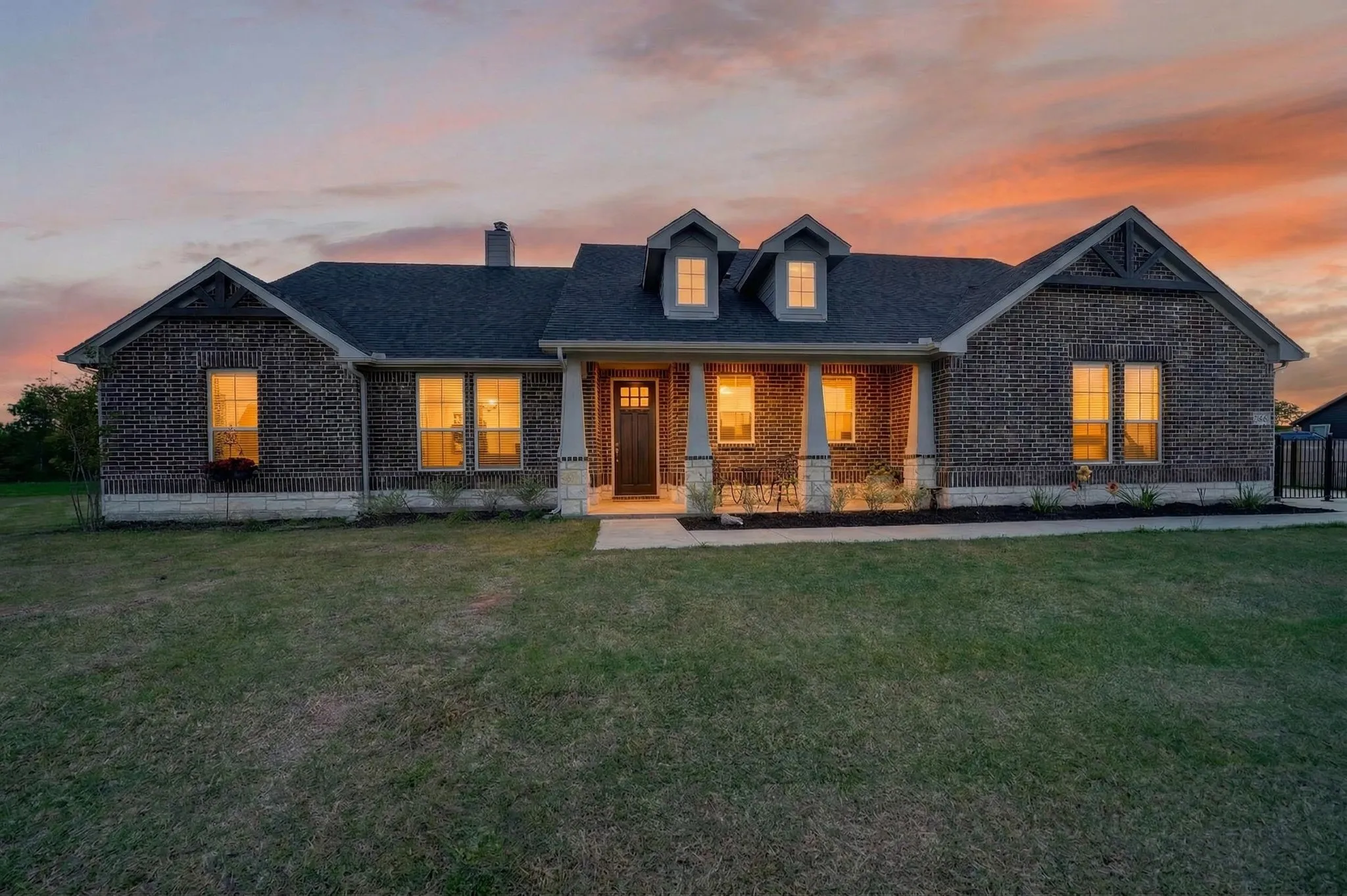 Brick facade residence featuring a covered front porch with stone columns, dormer windows, a dark shingle roof, and an expansive front lawn