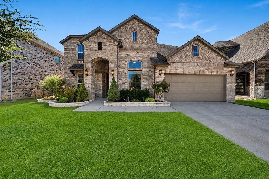 Brick exterior featuring arched entry, decorative stone accents, mature landscaping, an attached garage, and a concrete driveway