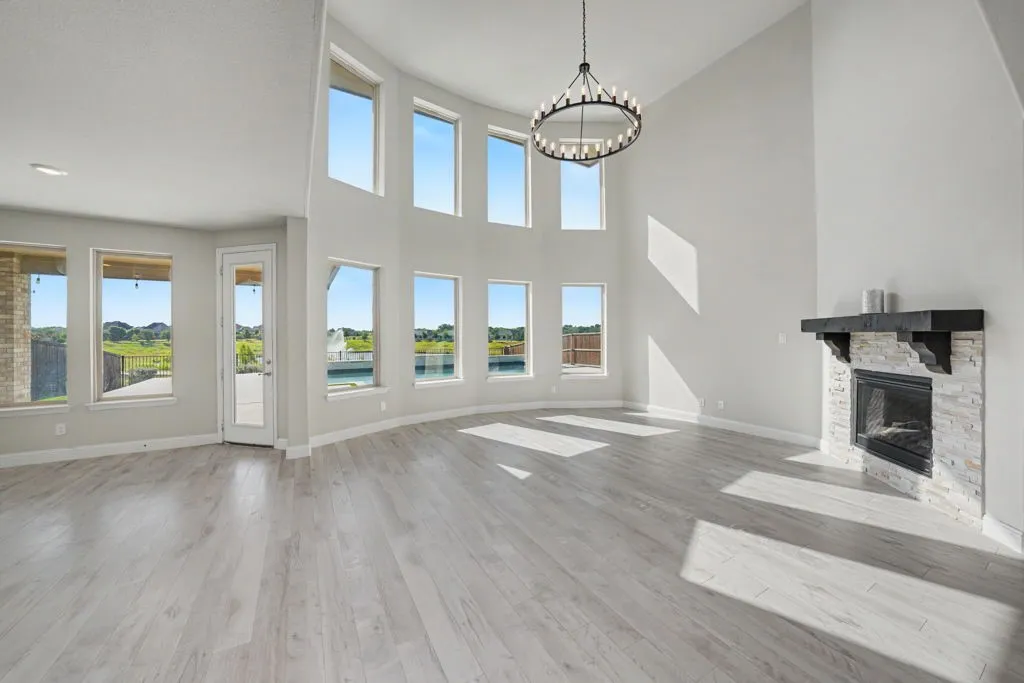 Two-story great room with abundant windows, wood-finish flooring, a stone-clad fireplace with a dark wood mantel, and a multi-tiered chandelier