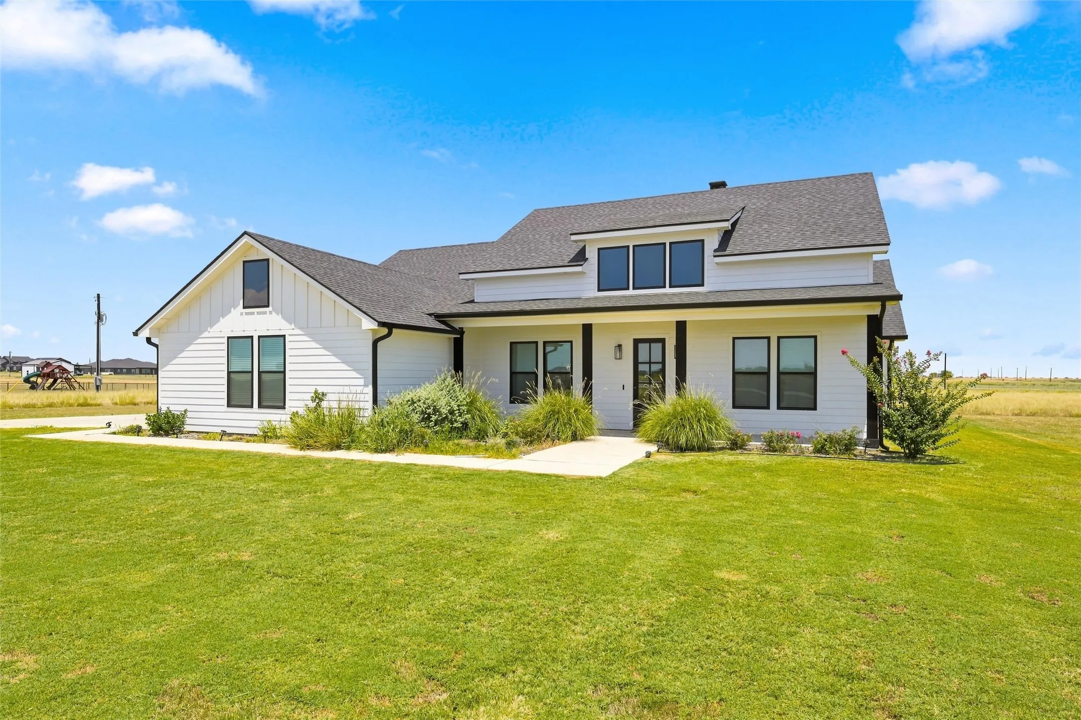Modern farmhouse exterior featuring white board and batten siding, a black shingle roof, black-trimmed windows, a covered front porch, and a concrete walkway