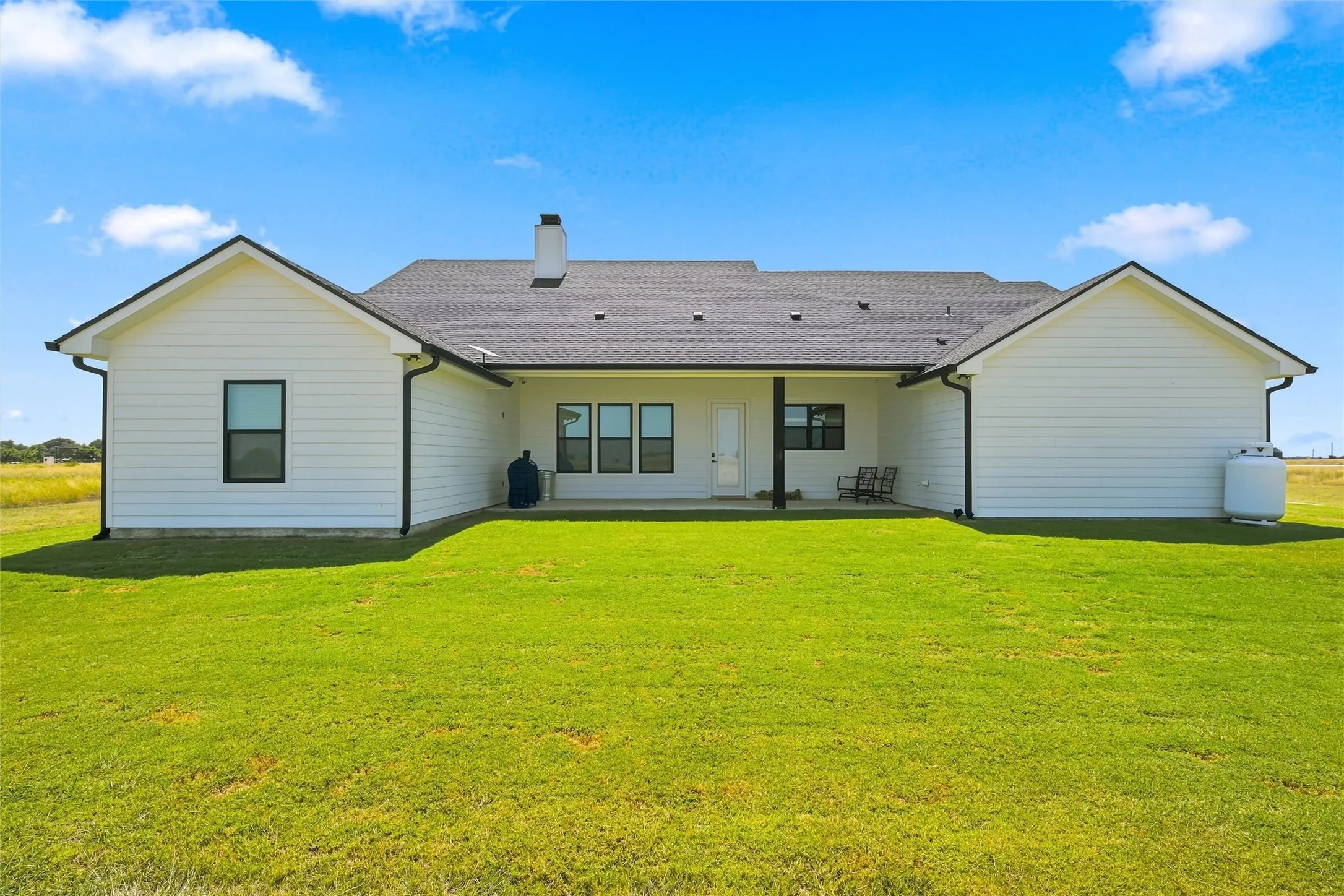 Rear exterior featuring white horizontal siding, a dark asphalt shingle roof, black window frames, a covered patio area, and a substantial green lawn