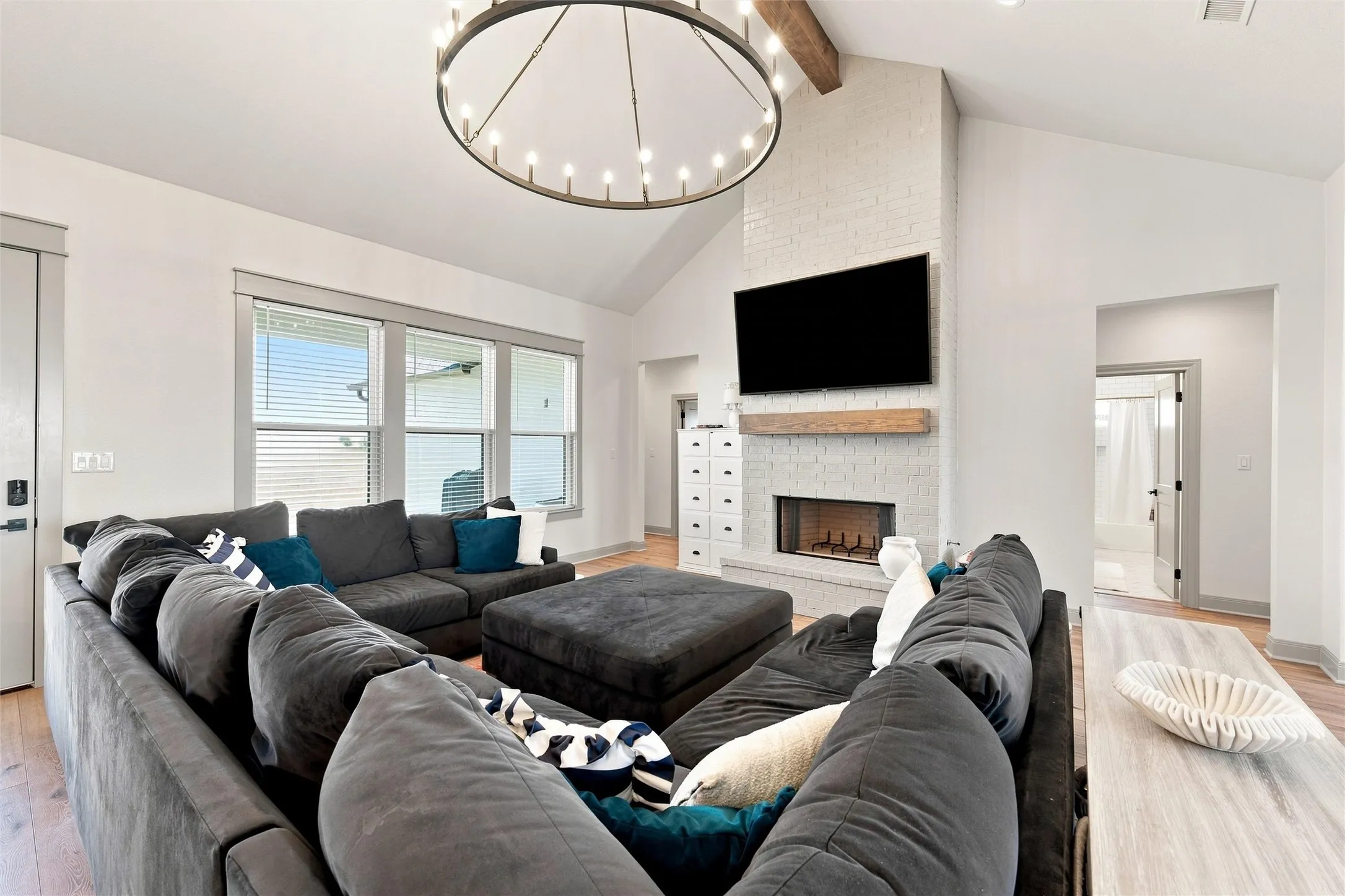 Living area with vaulted ceiling, exposed wood beam, large chandelier, and a white brick fireplace with a wood mantle