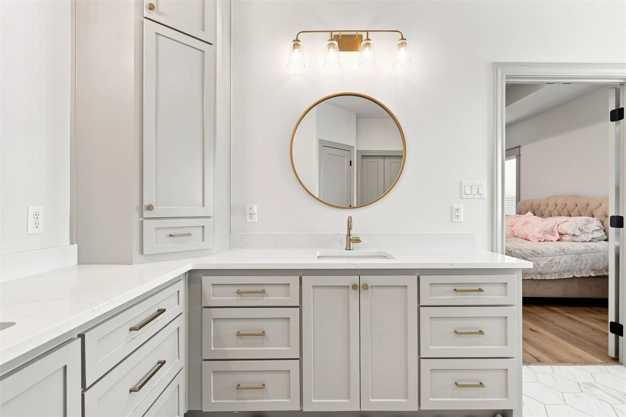 Bathroom vanity featuring light gray cabinetry, white countertops, a single basin sink, a circular mirror with a gold-tone frame, and a four-light fixture with a gold-tone finish