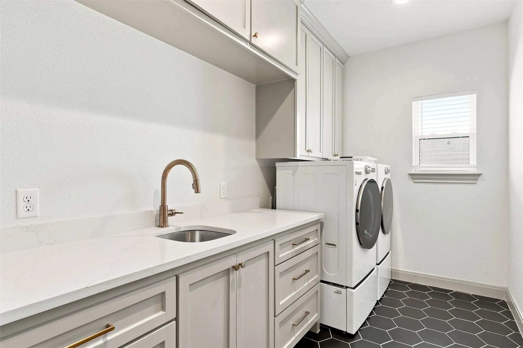 Utility room featuring a built-in stainless steel sink with a brushed brass faucet, white quartz countertop, light gray cabinetry with gold-finish hardware, and dark hexagonal floor tiling
