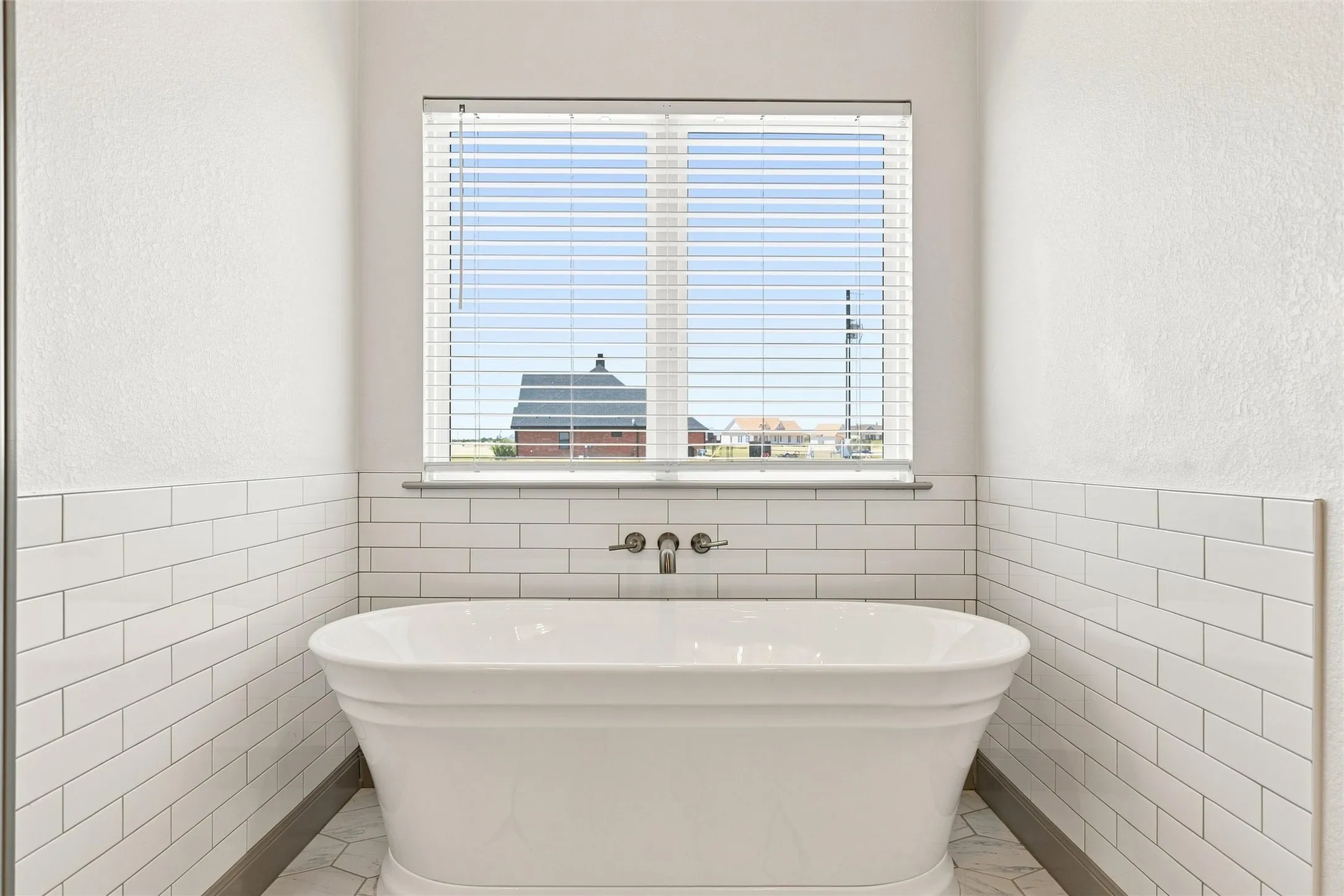 Freestanding soaking tub with floor-mounted fixtures, subway tile wainscoting, and a window with horizontal blinds