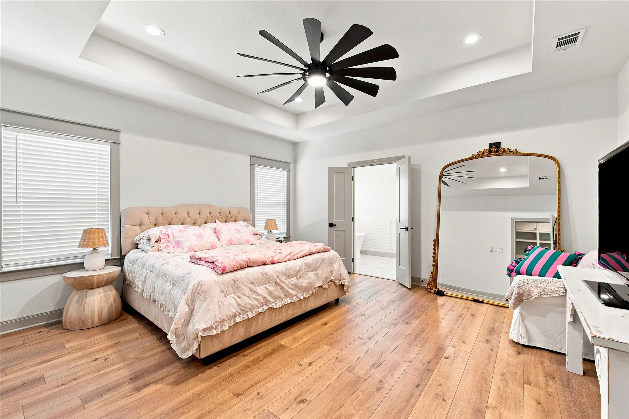 Spacious primary bedroom featuring a tufted upholstered bed with wood-finish flooring, recessed ceiling with modern black ceiling fan, large arched mirror with wood frame, and abundant natural light from multiple windows with horizontal blinds