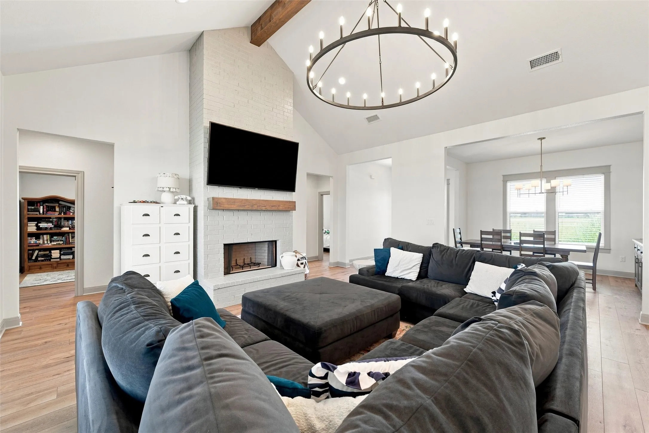 Living area featuring a floor-to-ceiling white brick fireplace with a wood mantle, vaulted ceilings with wood beams, and a large circular chandelier