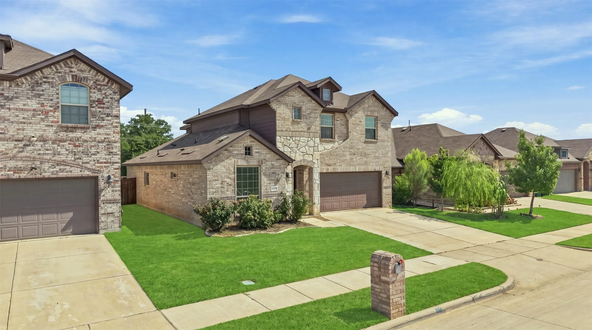 Two-story residence featuring a brick and stone facade, an attached two-car garage, manicured front lawn, a paved driveway, and mature landscaping