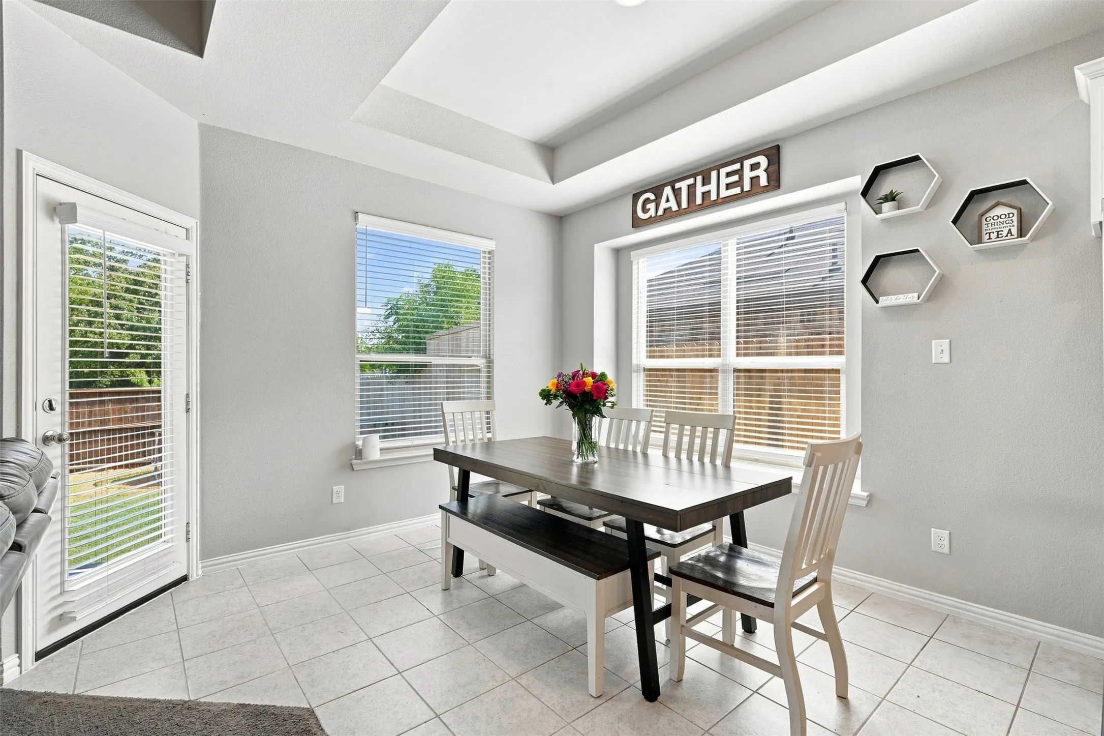 Bright dining area featuring a tray ceiling, tile flooring, two large windows with blinds, and a glass-paneled exterior door