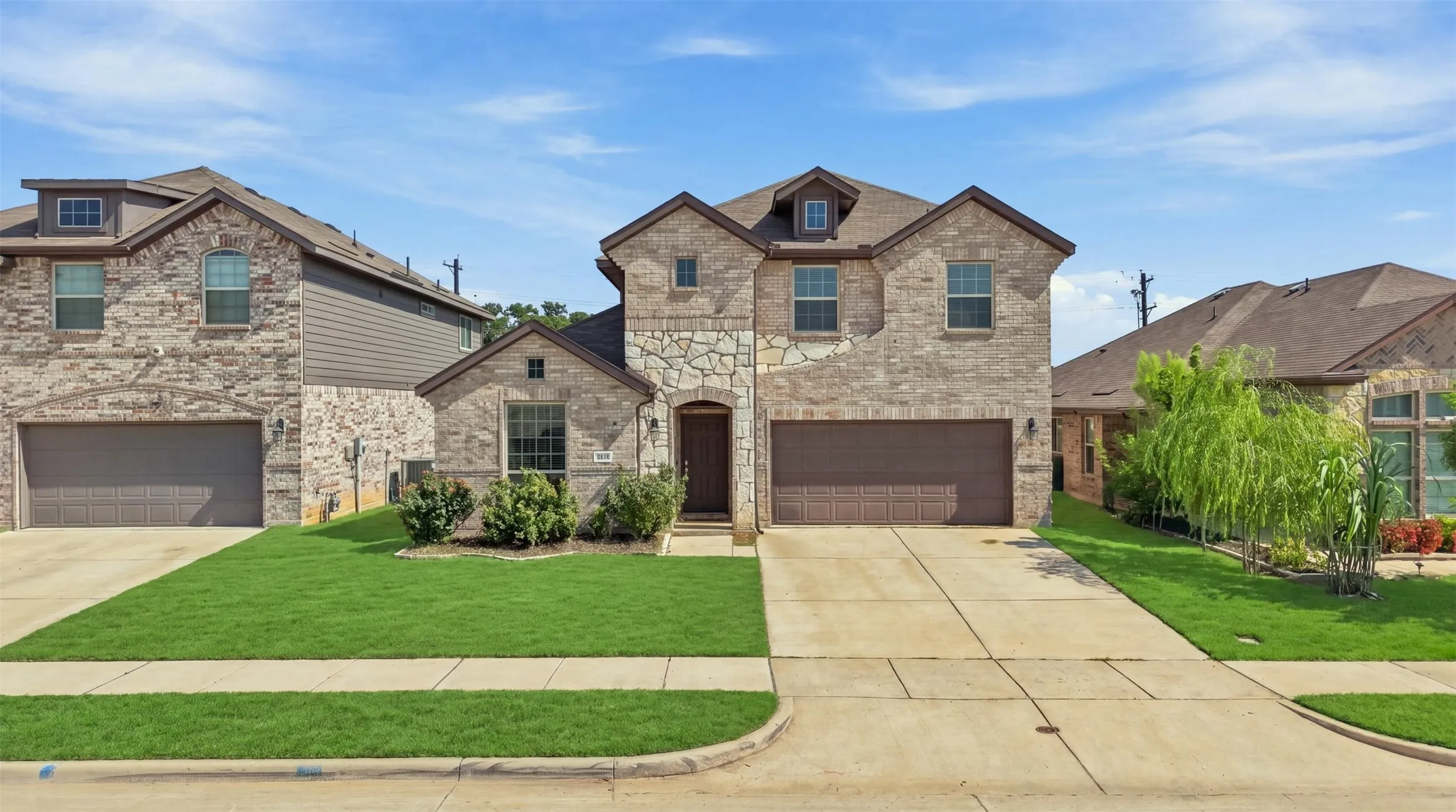 Two-story stone and brick facade residence featuring a two-car garage, dormer windows, and a landscaped front yard with a concrete driveway