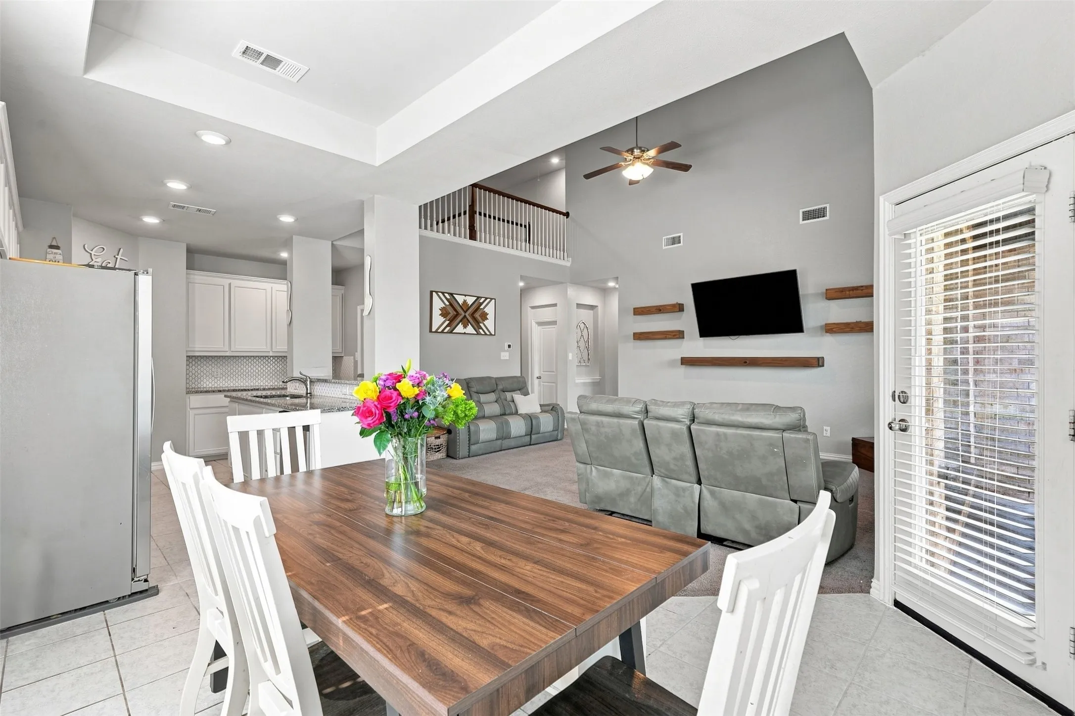 Open concept living space featuring a wood-finish dining table, light tile flooring, and a kitchen with white cabinetry and a mosaic tile backsplash