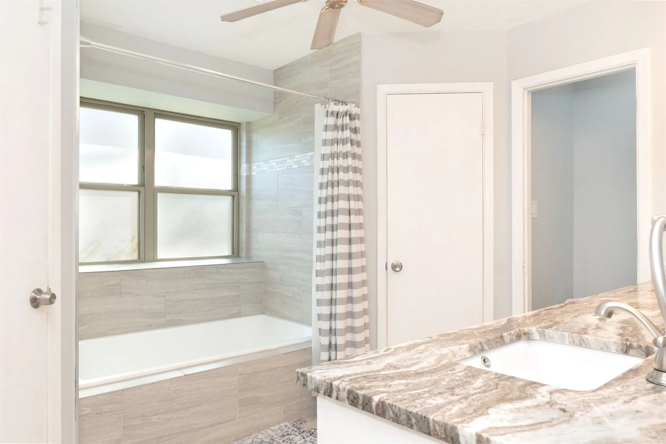 Bathroom featuring a tiled tub surround with an integrated window, a built-in bathtub, a vanity with an undermount sink, a stone-finish countertop, and an overhead ceiling fan