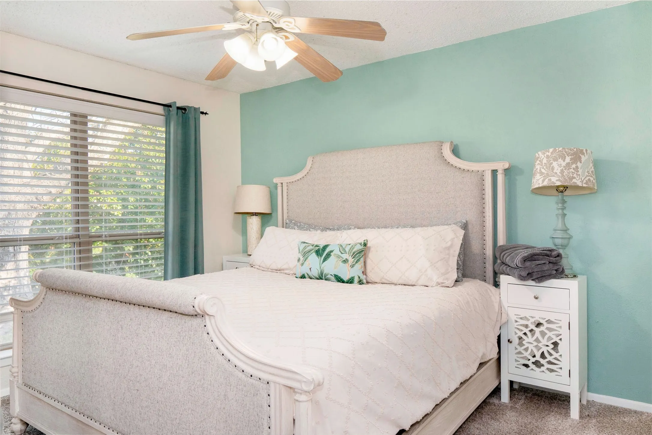 Spacious bedroom featuring light-colored carpeting, a ceiling fan with light fixture, and a large window with horizontal blinds and teal curtains