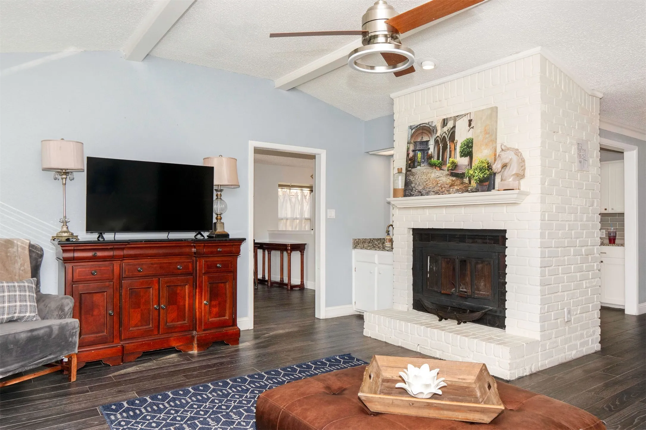 Living area featuring a painted brick fireplace, wood-finish flooring, vaulted ceilings with exposed beams, and a ceiling fan