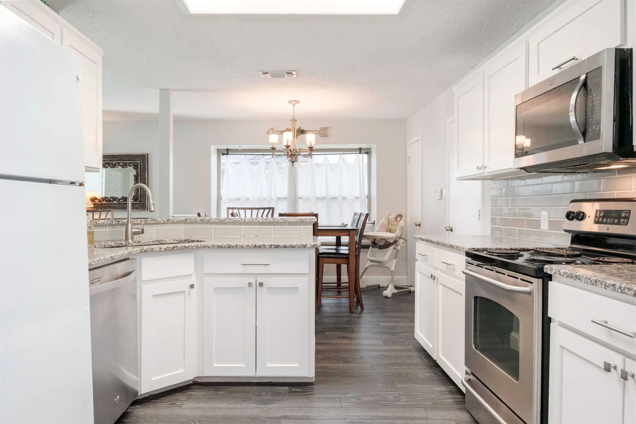 Bright, galley-style kitchen featuring white cabinetry, stainless steel appliances including a gas range and built-in microwave, granite countertops, subway tile backsplash, and an island with seating