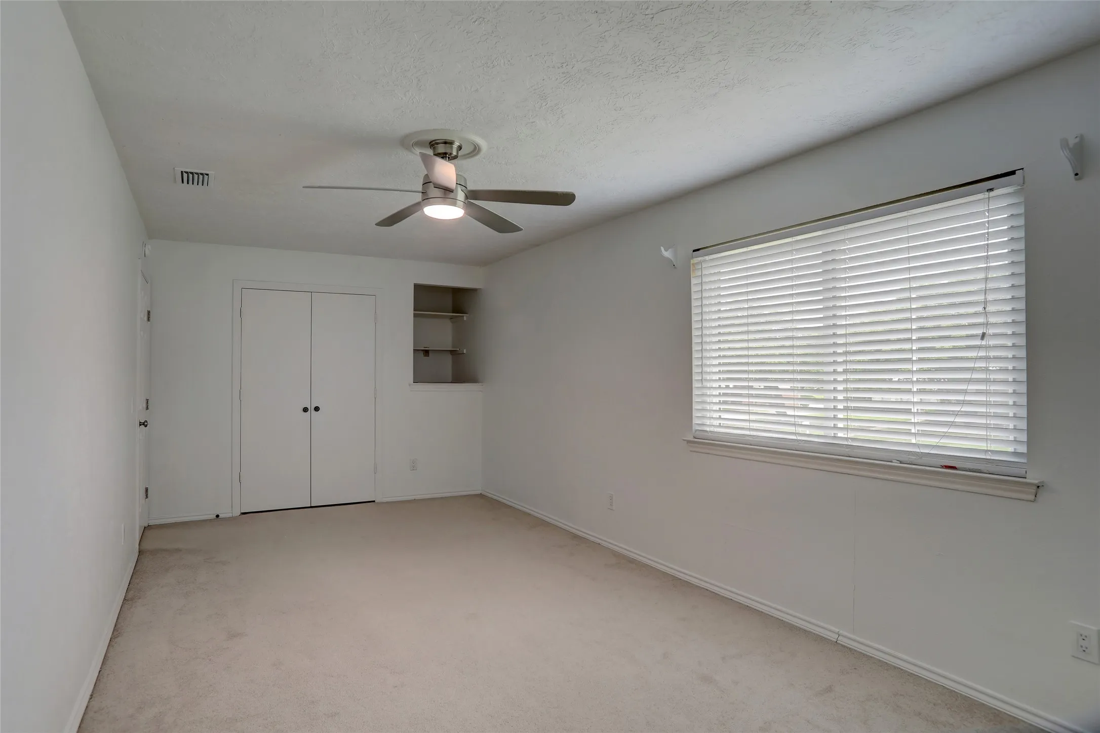 Spacious bedroom featuring neutral carpeting, ceiling fan with integrated lighting, and abundant natural light from a large window with horizontal blinds