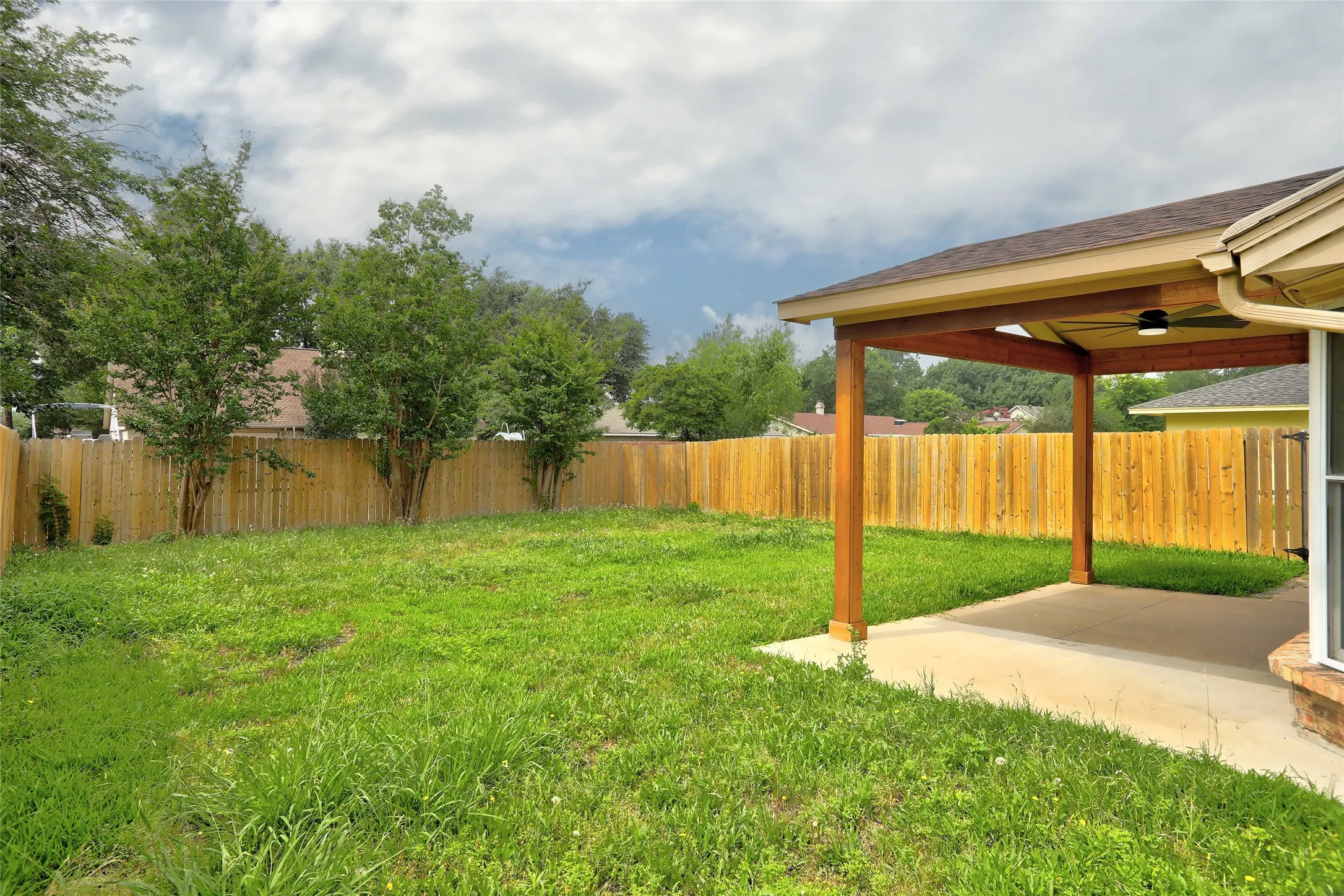 Expansive backyard featuring a covered patio with a ceiling fan, a full privacy fence, and mature trees along the perimeter