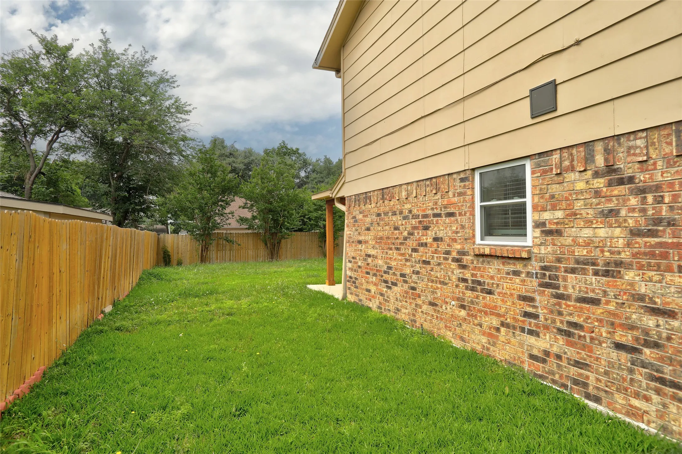 Spacious side yard featuring a new privacy fence, mature trees, a manicured lawn, and brick and siding exterior