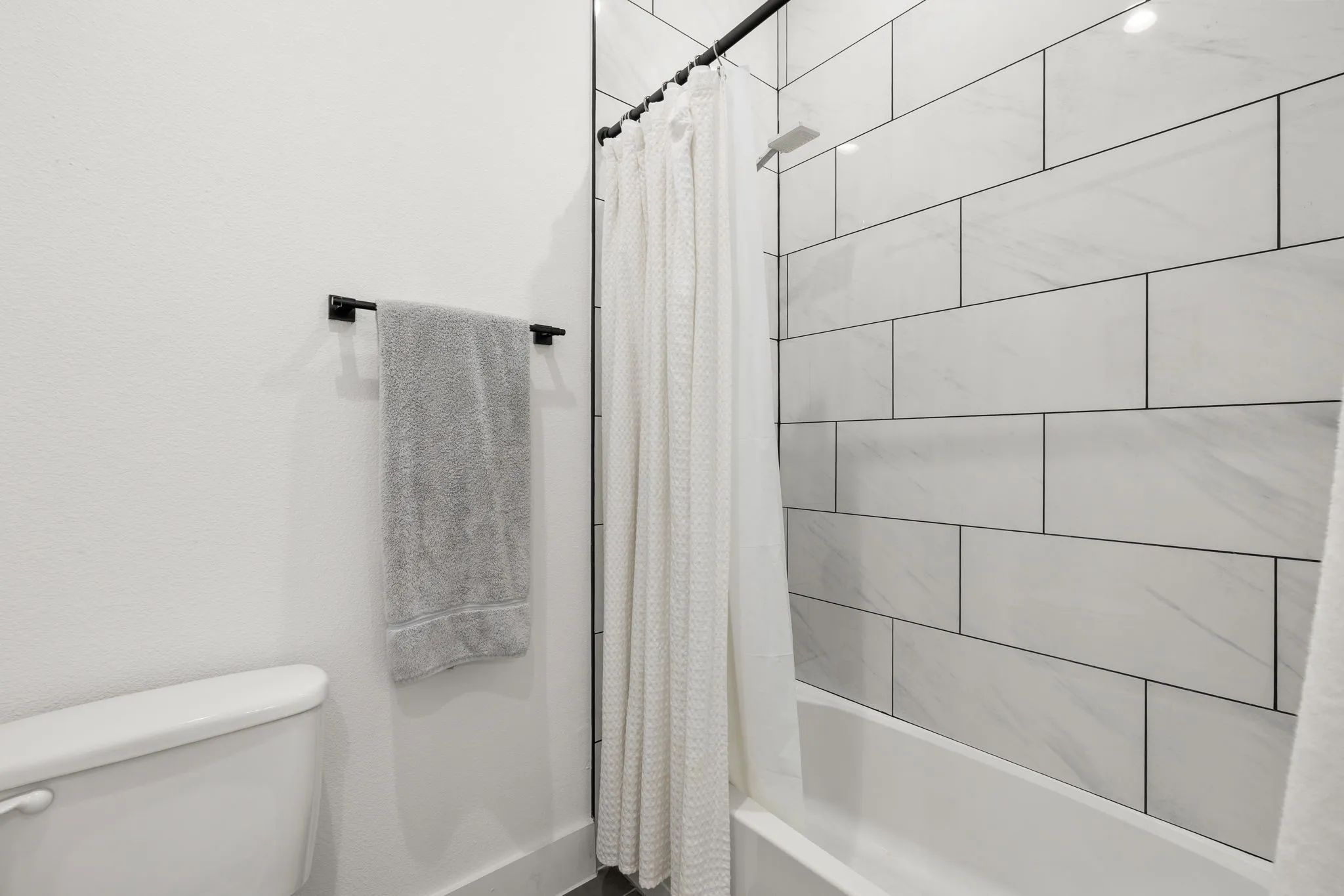 Bathtub and shower combination featuring white subway tile surround with dark grout, a matte black shower rod, and a white porcelain toilet