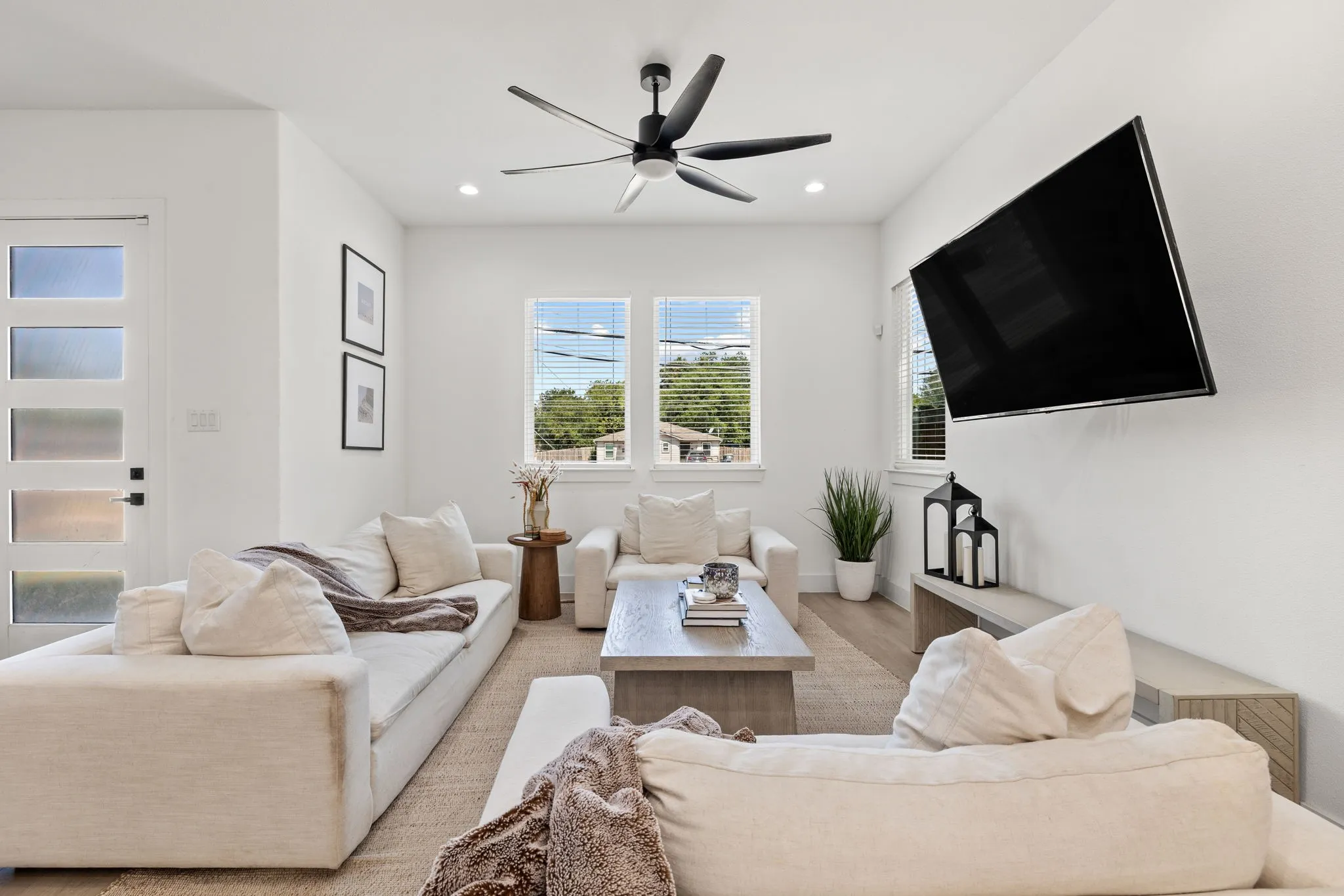 Living area featuring wood-finish flooring, two large windows with blinds, a ceiling fan with integrated lighting, recessed lighting, and a modern paneled door with frosted glass inserts