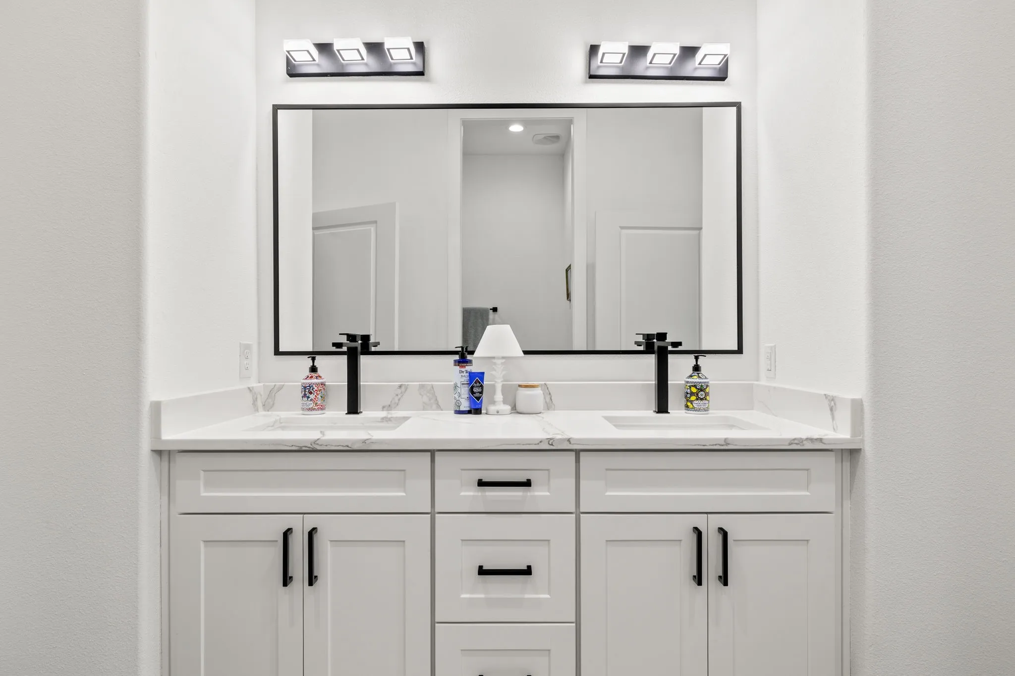 Spa-inspired dual-vanity bathroom featuring a floating white cabinetry base with ample storage, quartz countertop, matte black faucets and hardware, and a large framed mirror with integrated lighting