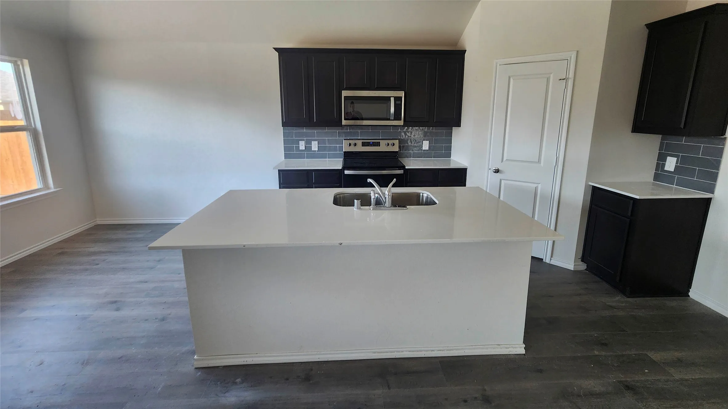Spacious kitchen featuring a large quartz island with waterfall edge, stainless steel appliances including built-in microwave, black cabinetry with gray subway tile backsplash, dual-basin sink with chrome fixtures, and dark wood-finish flooring throughout the open layout