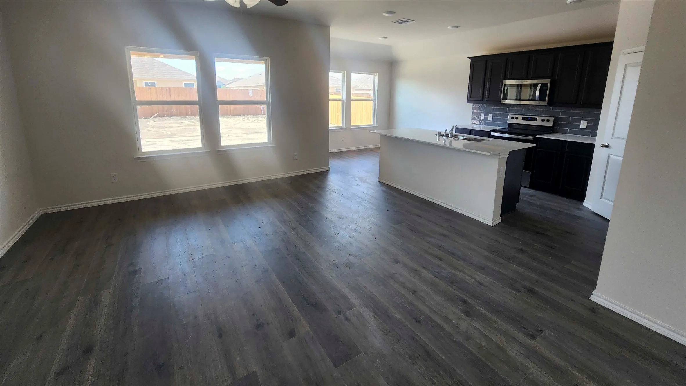 Open-concept living space featuring wood-finish flooring, a kitchen island with a sink, dark cabinetry, and a tile backsplash