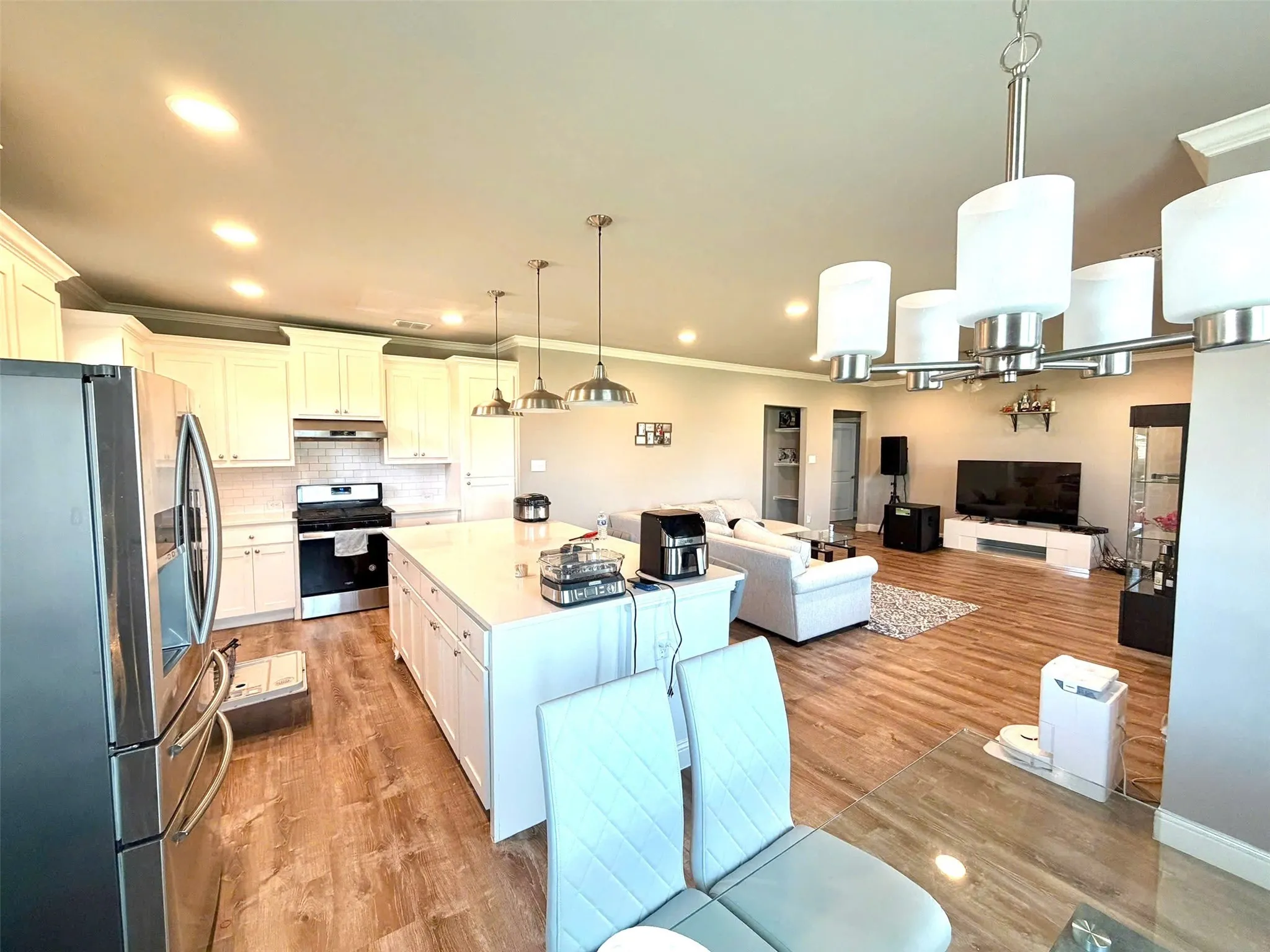 Open-concept living space featuring a central kitchen island with pendant lighting, white cabinetry, stainless steel appliances, and wood-finish flooring extending into the living area
