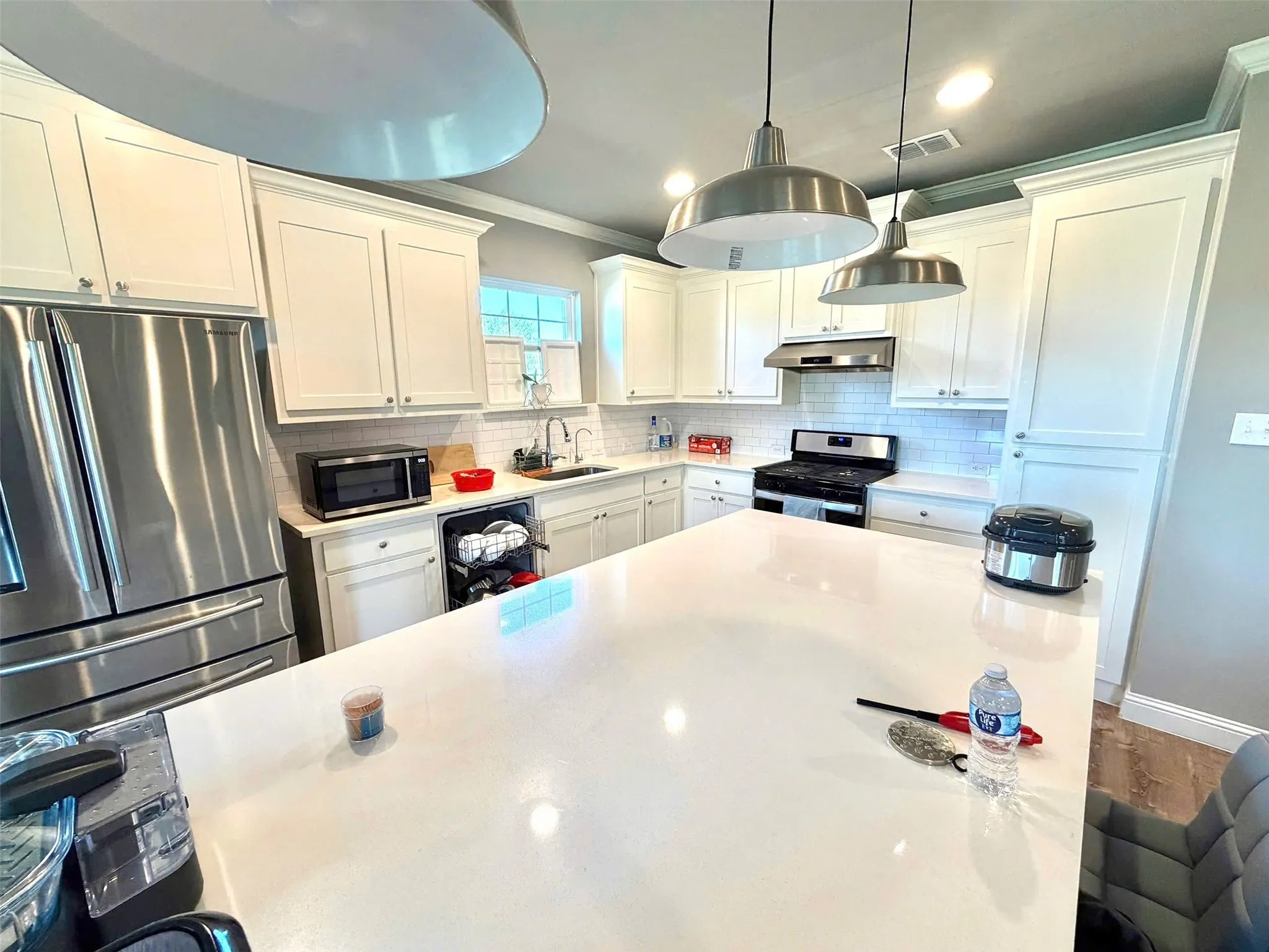 Kitchen featuring a large island with a light-toned countertop, stainless steel appliances, white cabinetry, subway tile backsplash, and industrial-style pendant lighting