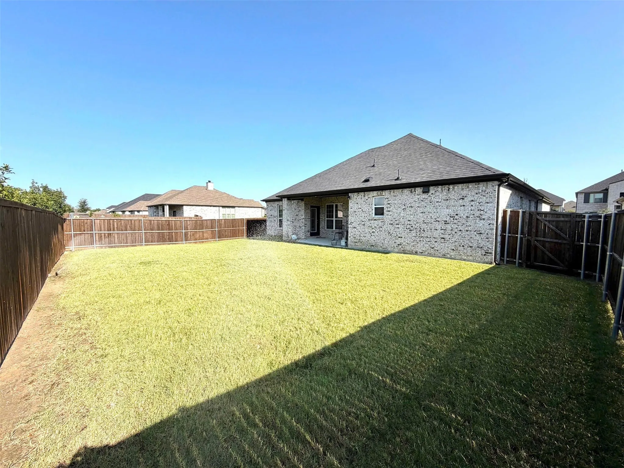 Spacious backyard featuring a large green lawn, wood privacy fencing, and a brick exterior with a covered patio