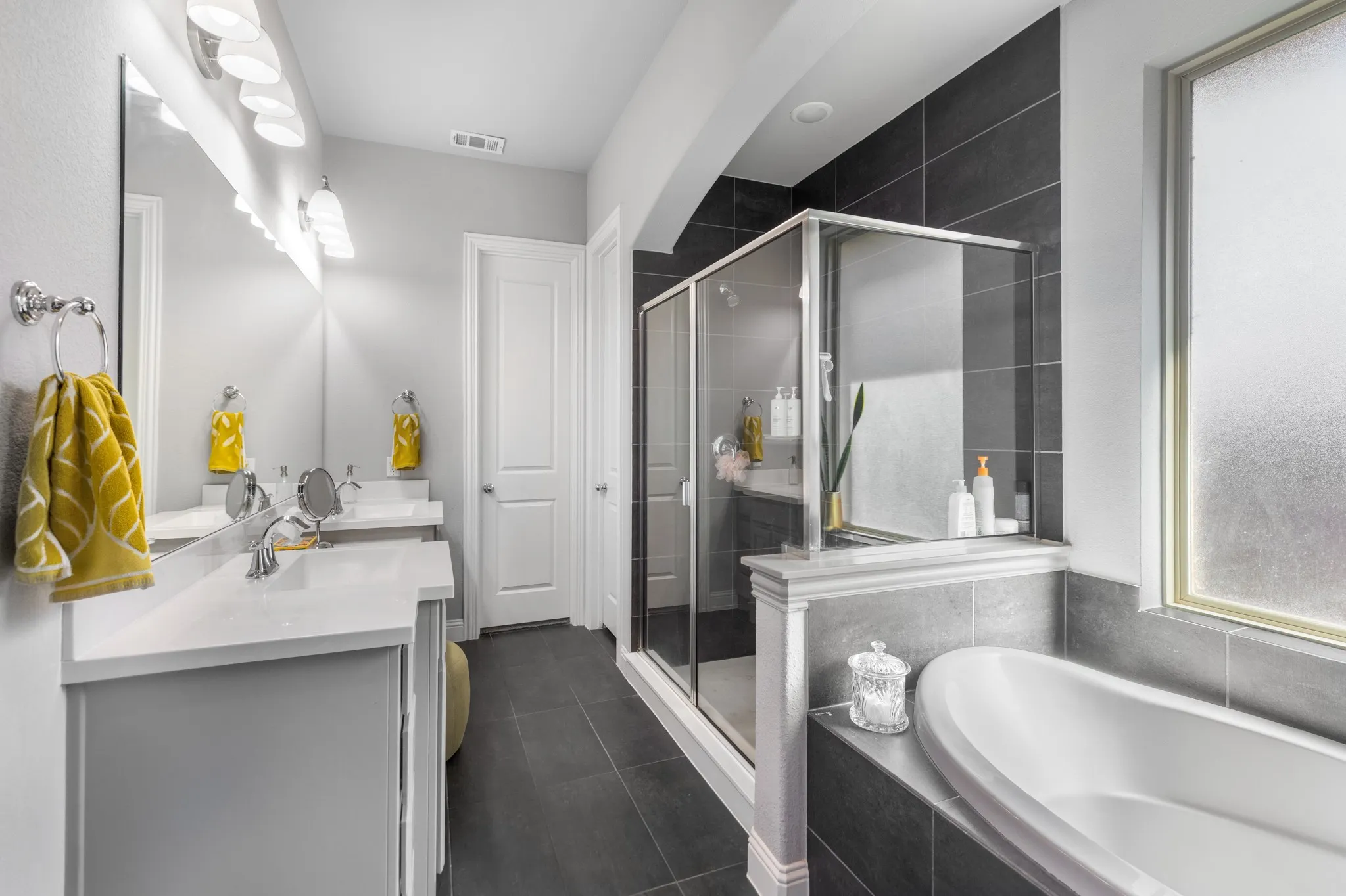 Spa-inspired primary bathroom featuring a frameless glass-enclosed shower with dark tile surround, freestanding soaking tub positioned beneath a frosted window, dual vanity with white countertop and integrated sink, chrome fixtures, and dark tile flooring throughout